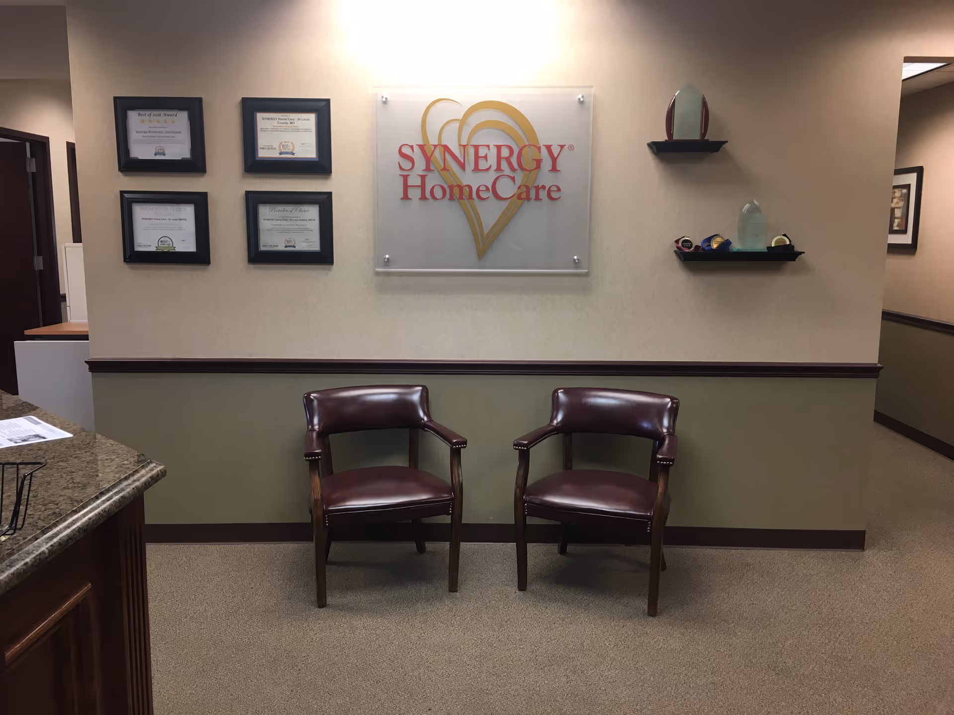 Two brown chairs sit in a reception area with Synergy HomeCare sign on wall.