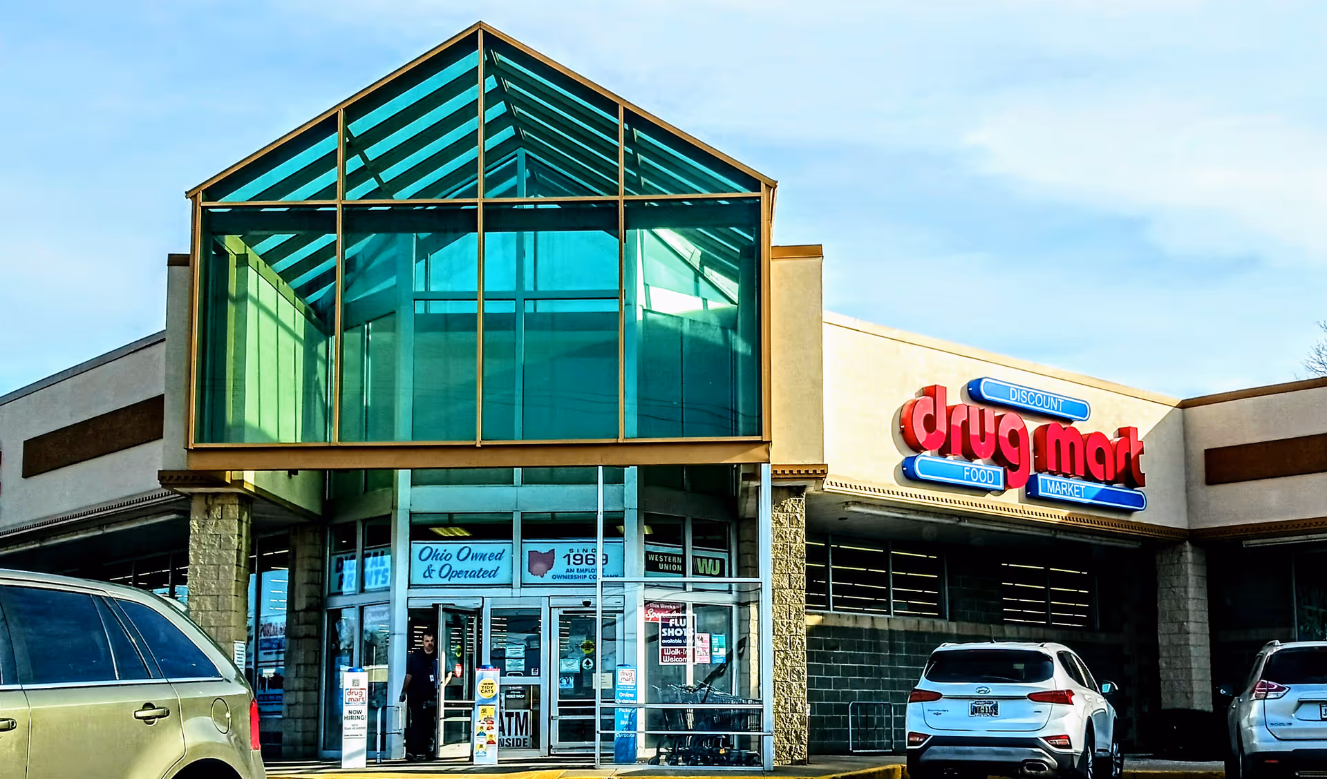 Front exterior of a Discount Drug Mart store with glass atrium entrance and parked cars