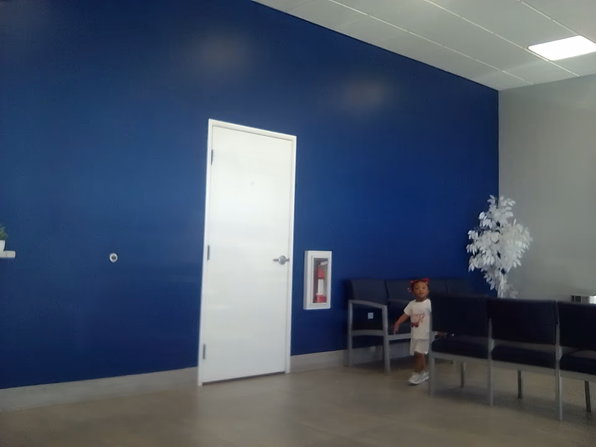 Interior lobby area with a blue wall, white door, row of chairs, and a child.
