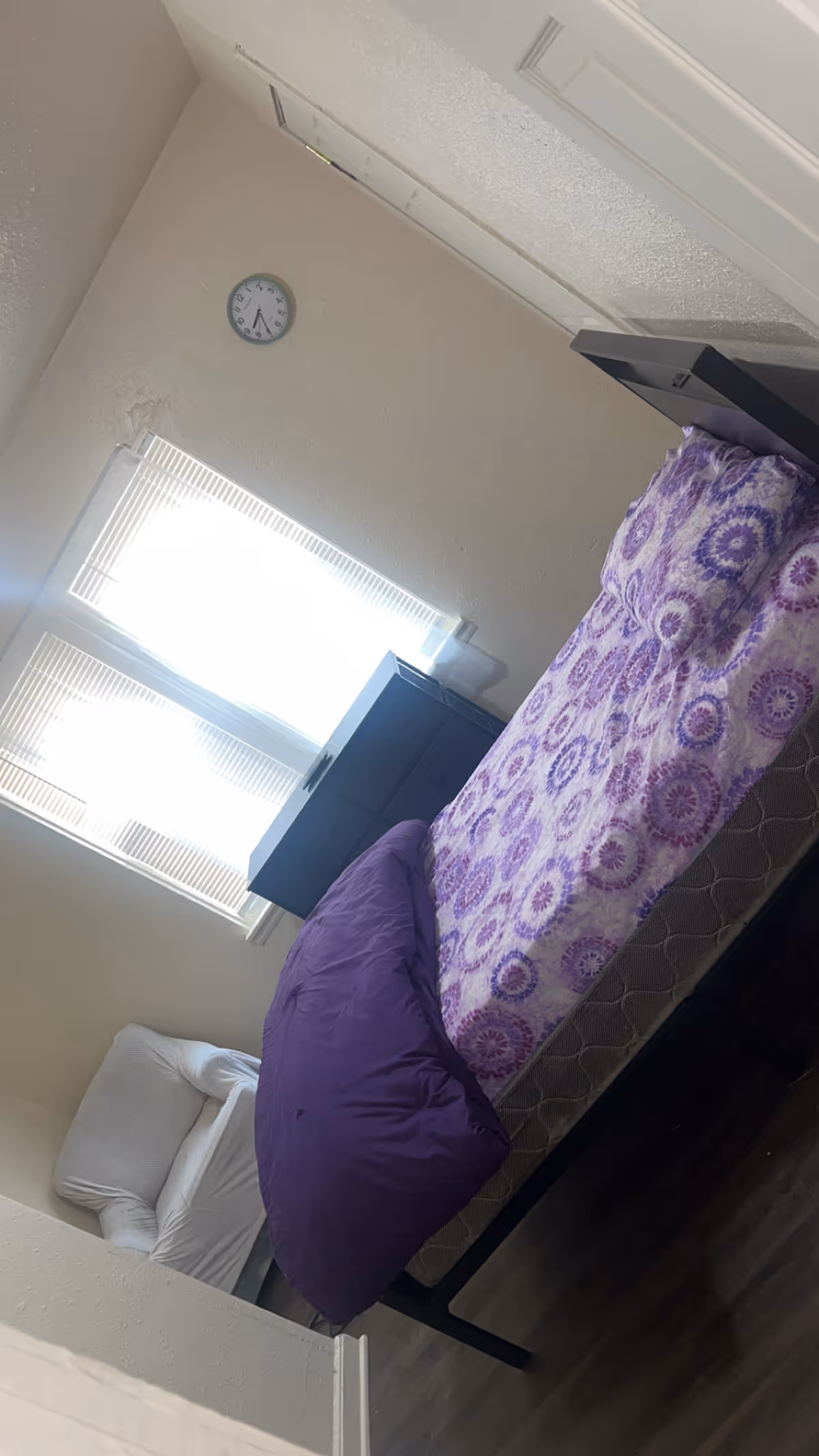 A small bedroom with a purple-patterned bed, window blinds, and a wall clock.