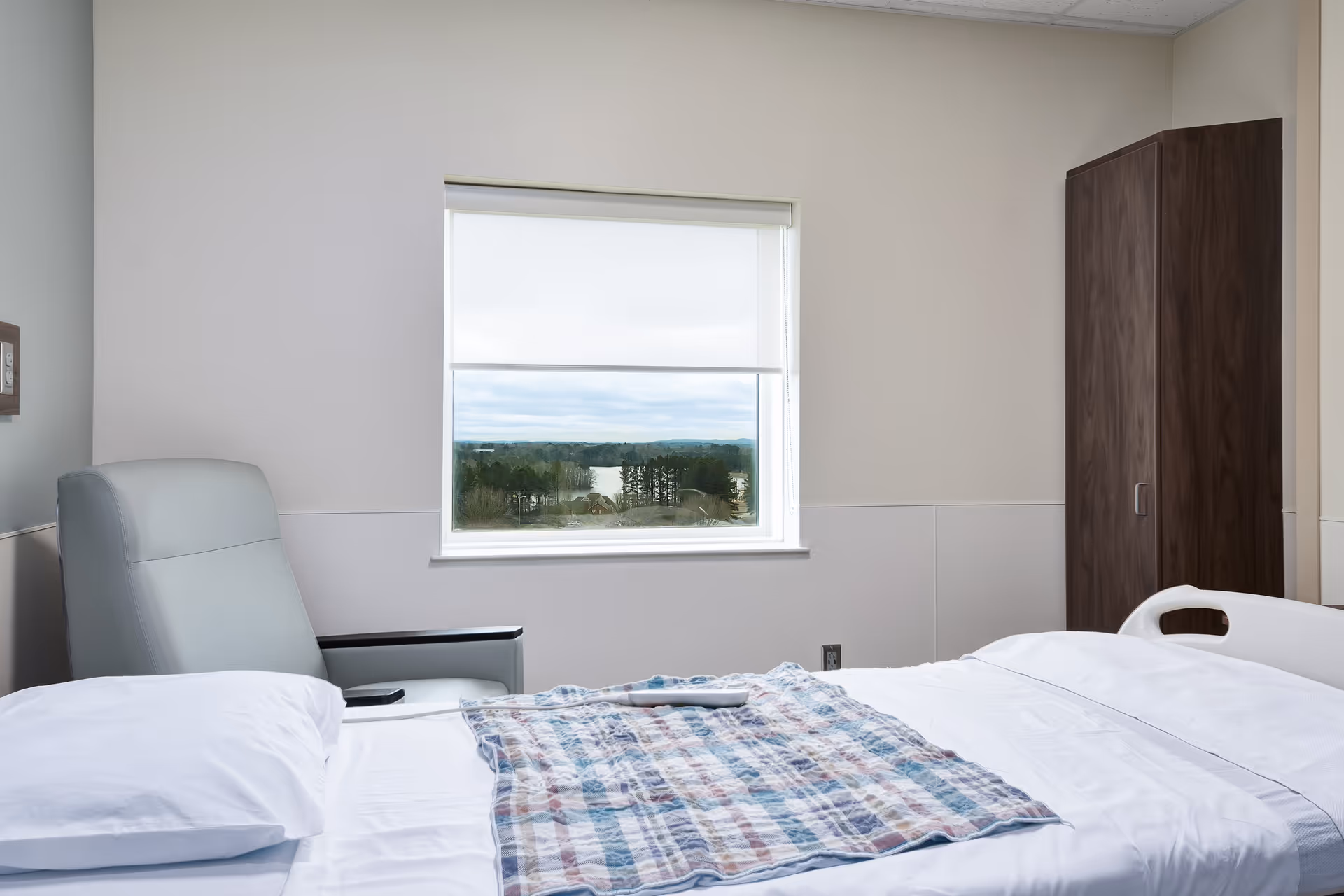 Hospital room interior with a bed, chair, wardrobe, and window view outside.