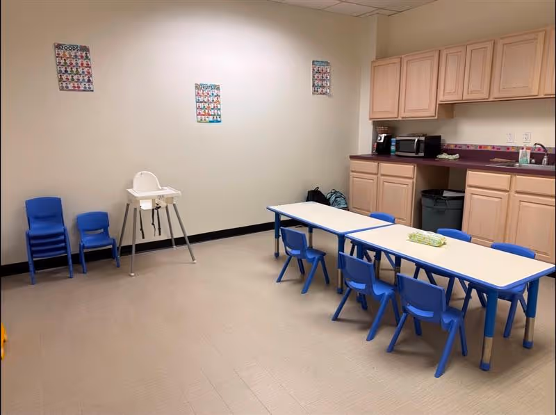 Activity room with child-sized blue chairs around a long table and kitchenette.