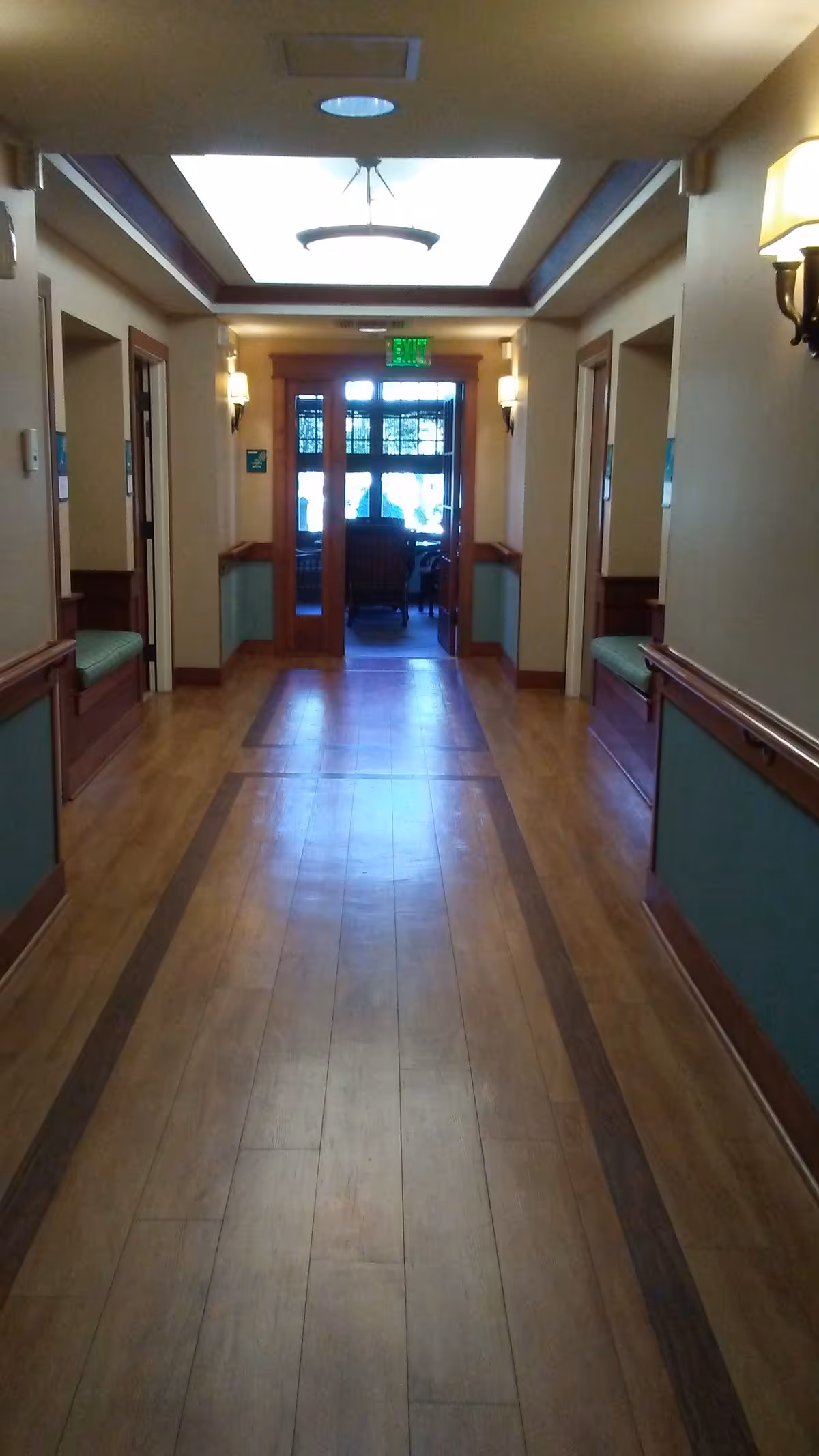 Hallway in a senior living facility with wooden floors, wall benches, and an exit at end.
