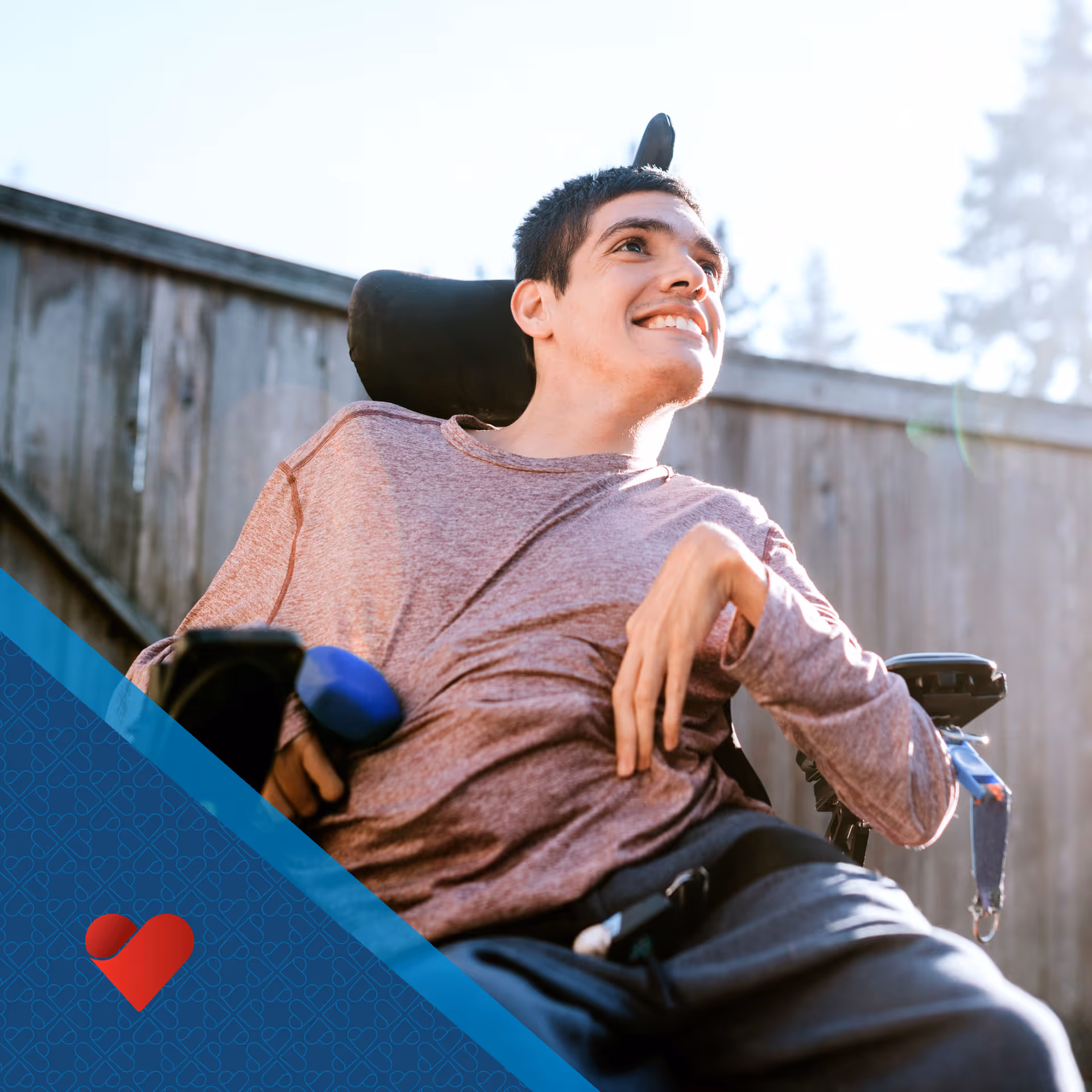Smiling person in a wheelchair outdoors beside a wooden fence.