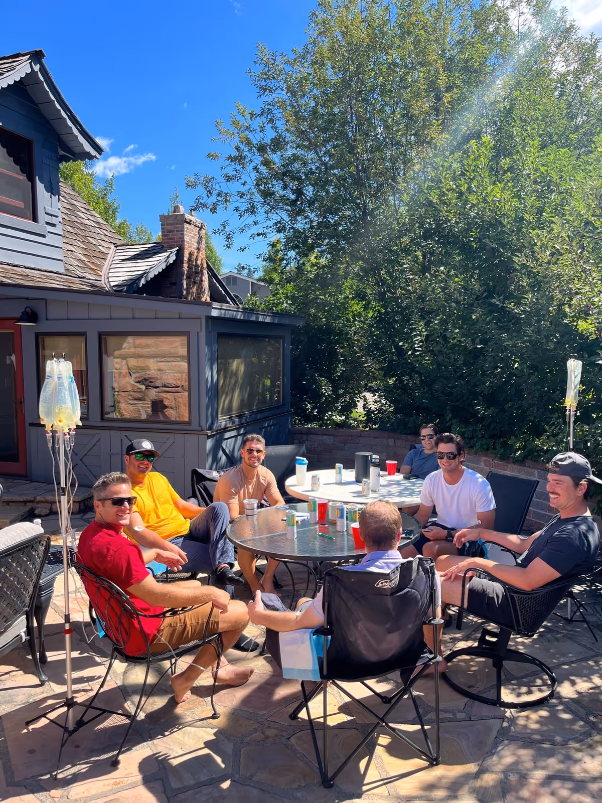 Group of adults sits around a round patio table outdoors beside a house.