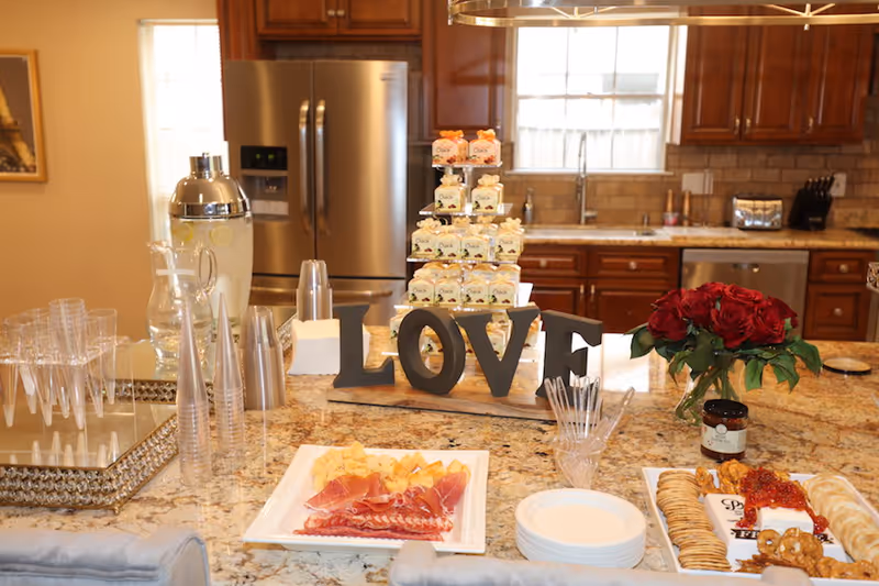A well-lit kitchen island displays a beverage dispenser, assorted foods, and a bouquet of roses.