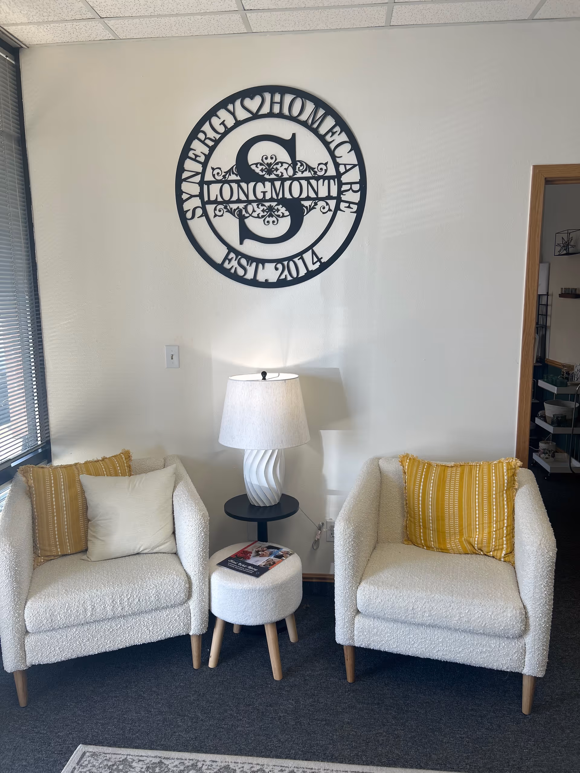 Two white armchairs with yellow pillows flank a small table and lamp under a logo.