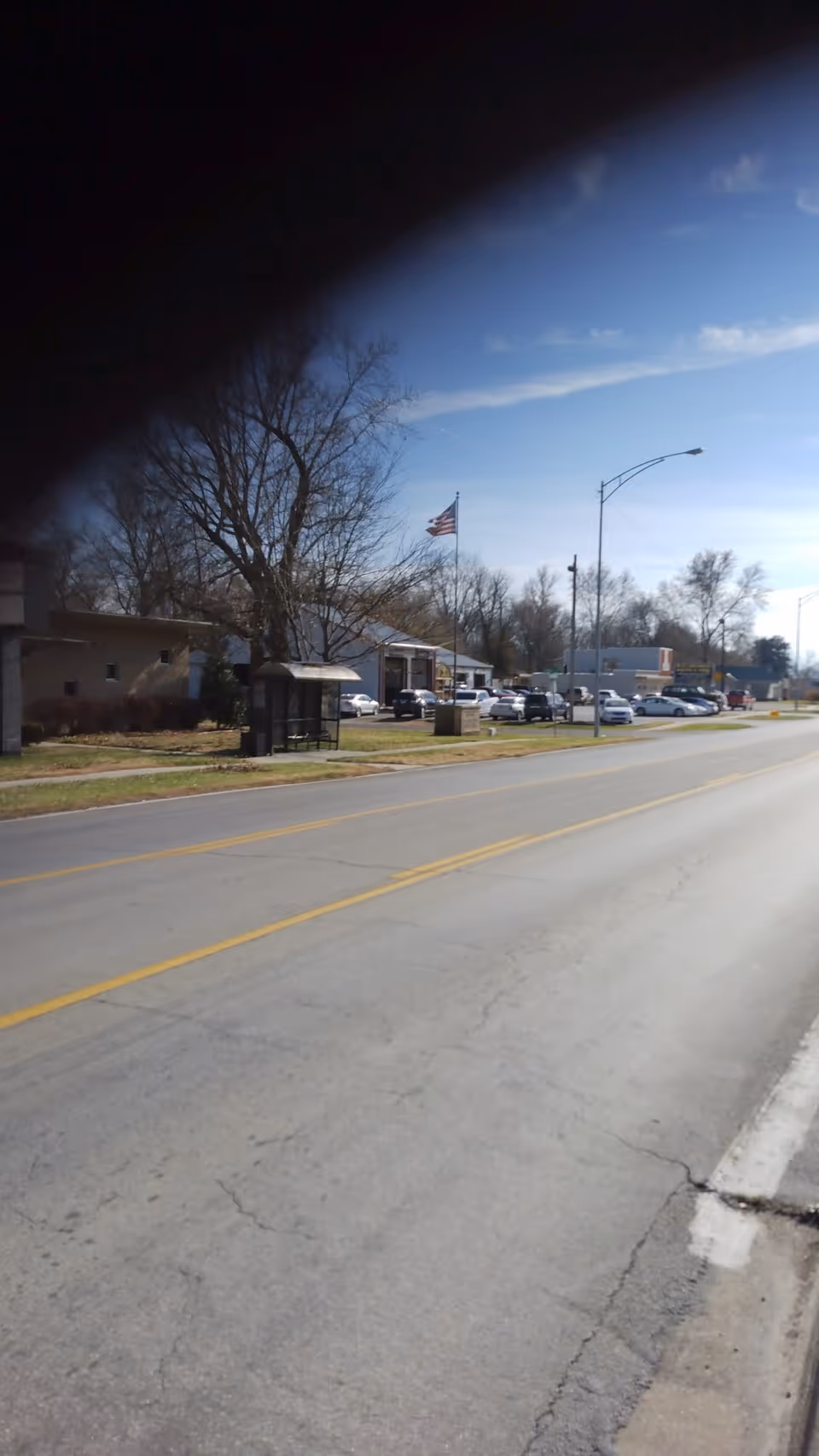 Exterior view of a facility along a road with parked cars, flag, and a bus shelter.