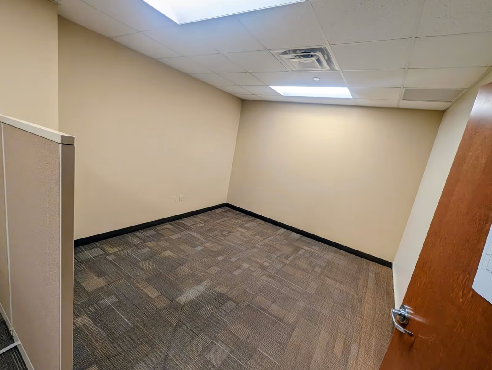 Empty beige office with carpet, drop ceiling tiles, and an open wooden door.