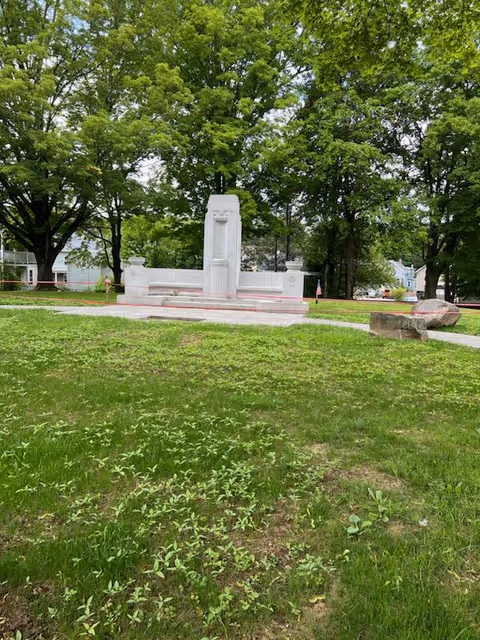 White monument centered in a grassy park with tall leafy trees surrounding it.