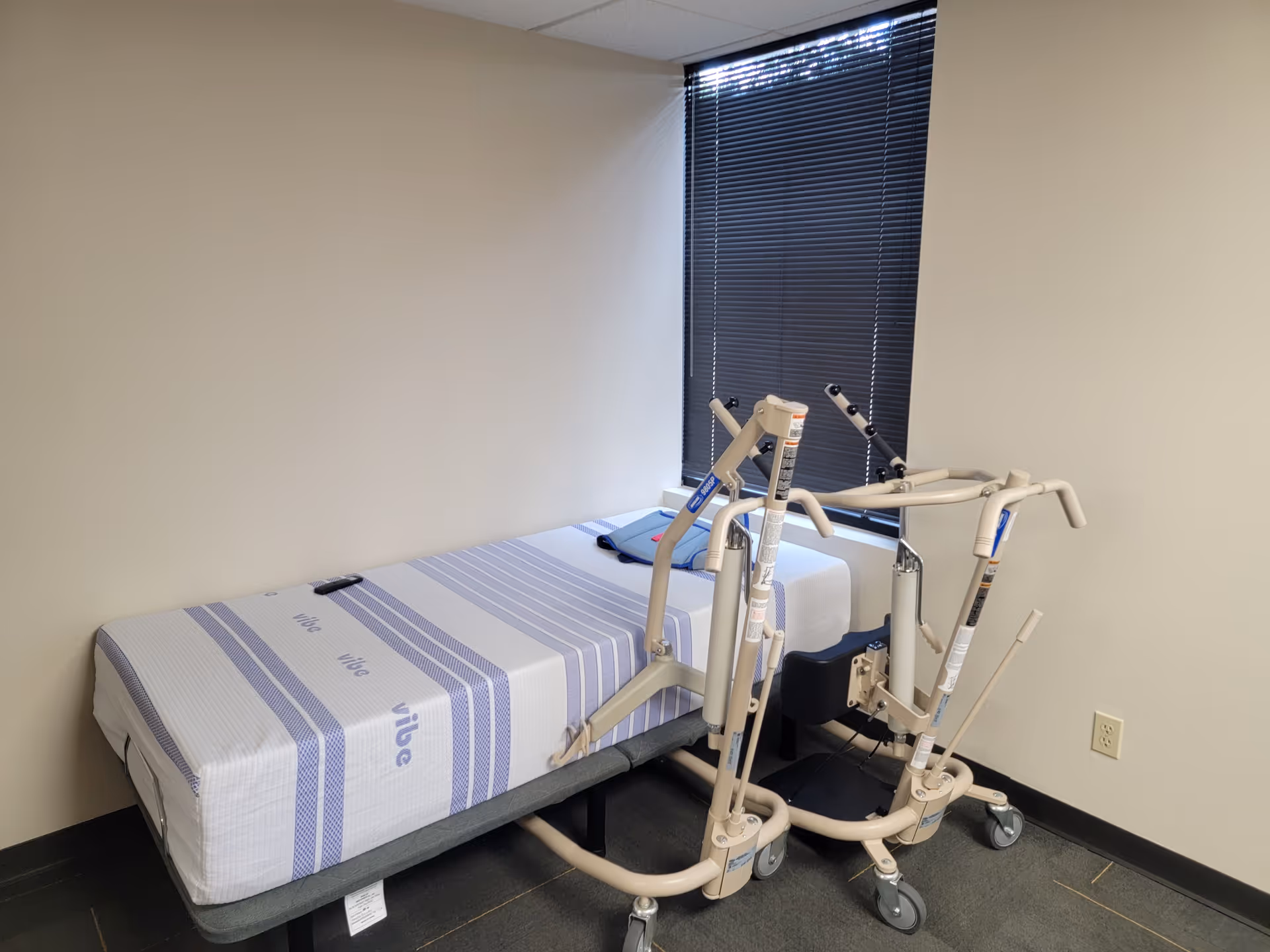 Small bedroom with a bed and patient lift device beside a window with blinds.