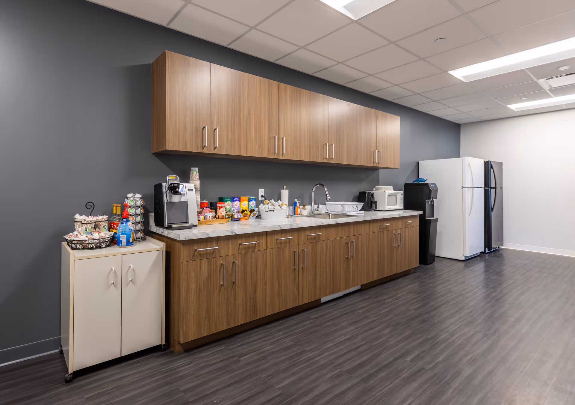 Long kitchenette with wood cabinets, appliances, and a refrigerator along a gray wall.