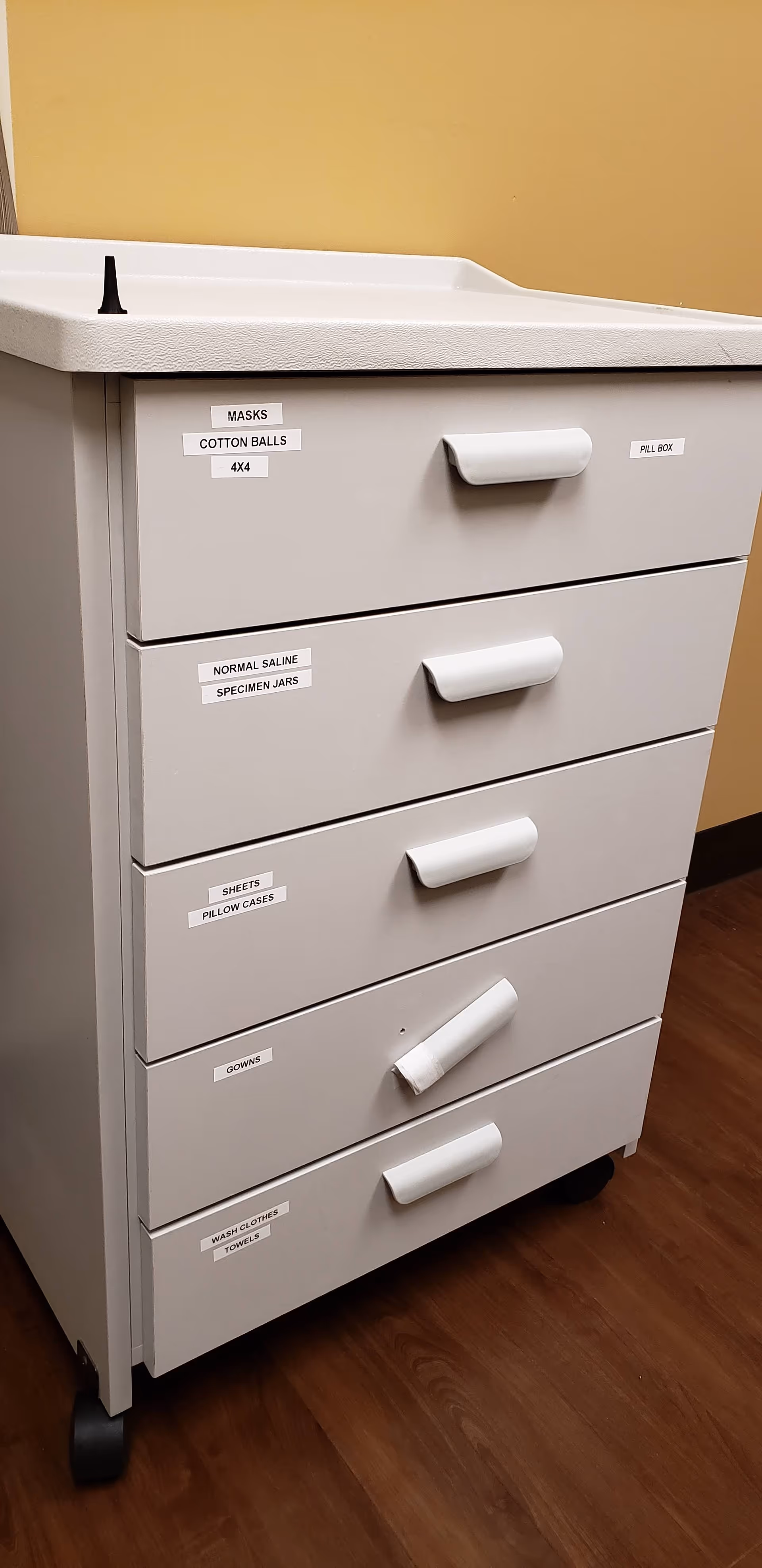 Gray medical cabinet with labeled drawers and a top tray sits beside a tan wall.