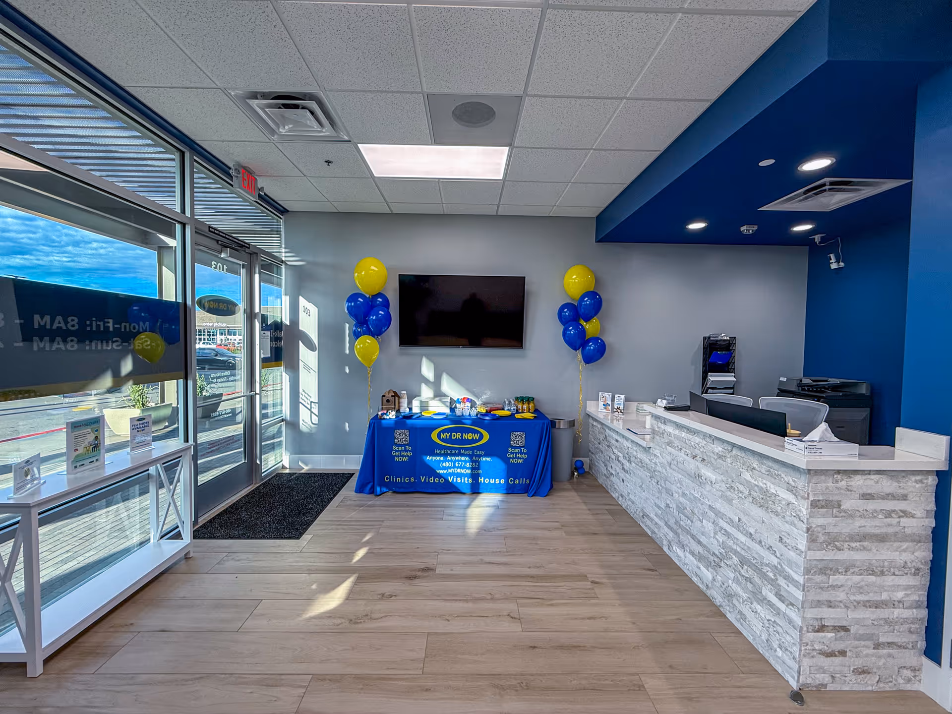Lobby reception area of MY DR NOW with a blue-accented desk, TV, and balloons.
