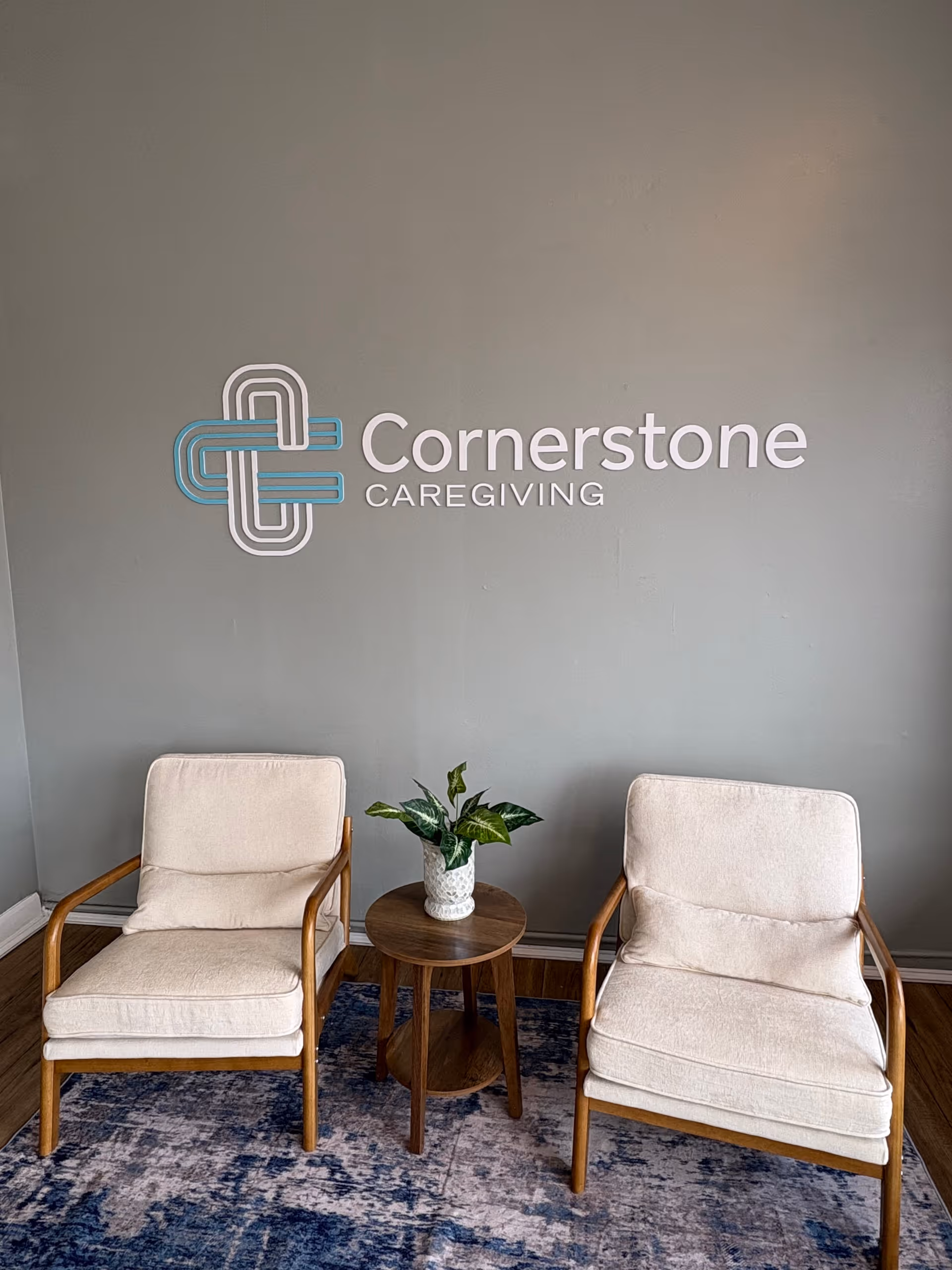 Two cream chairs flank a wooden table with a potted plant, in front of Cornerstone Caregiving.