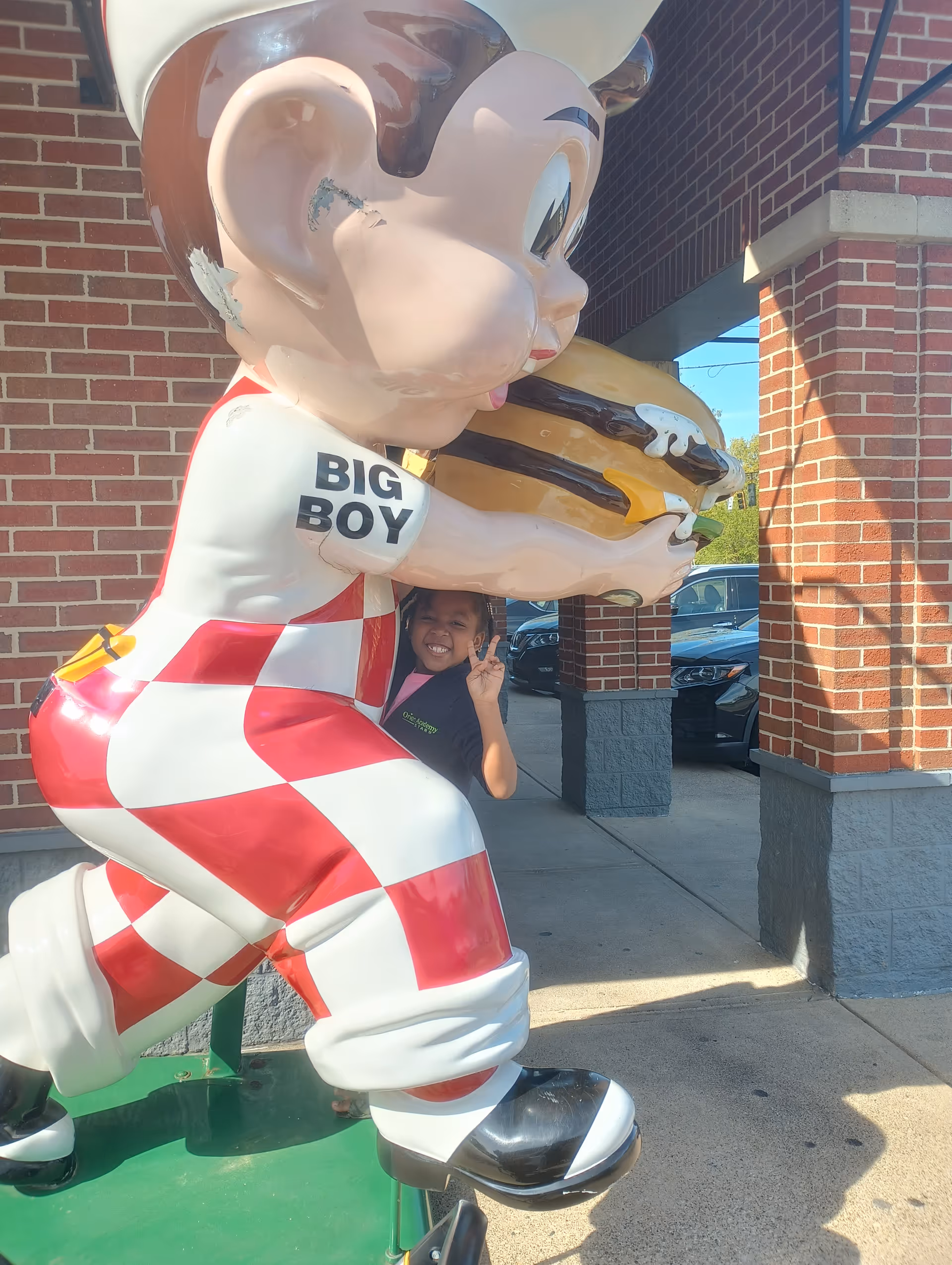 A large Big Boy statue outdoors beside a brick building, with a smiling child posing.