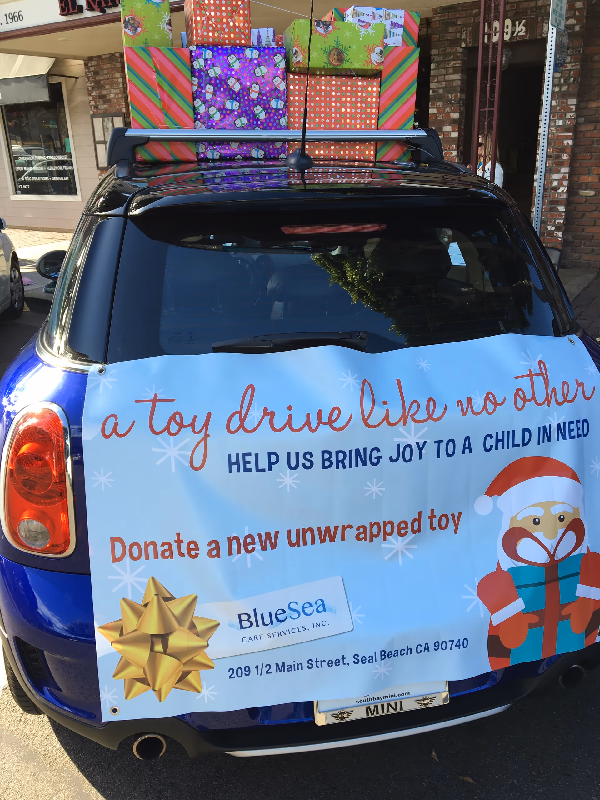 Rear of a blue car with a banner promoting a toy drive; storefront and gifts visible