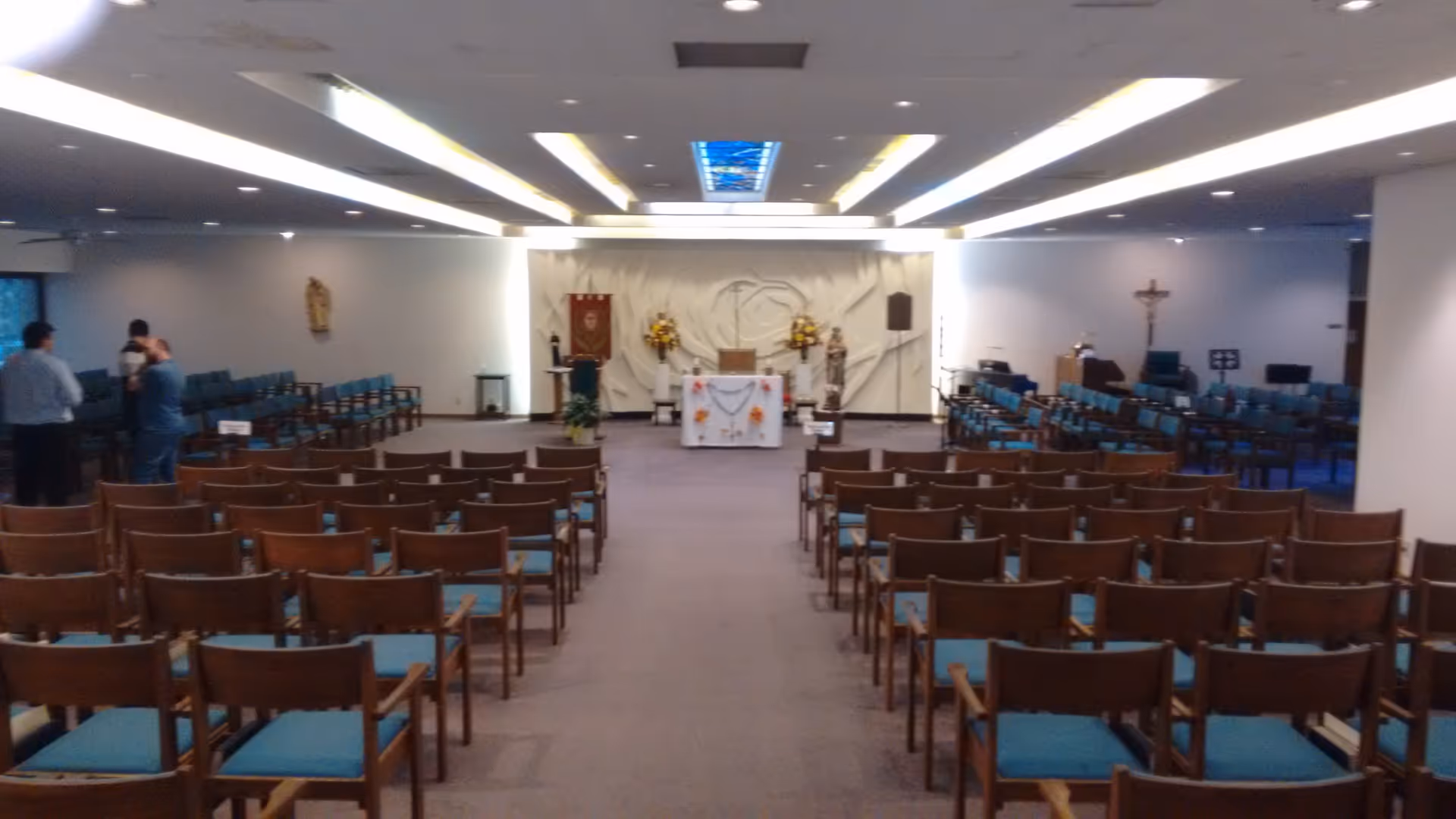 A spacious indoor chapel with rows of blue-cushioned chairs facing an altar.