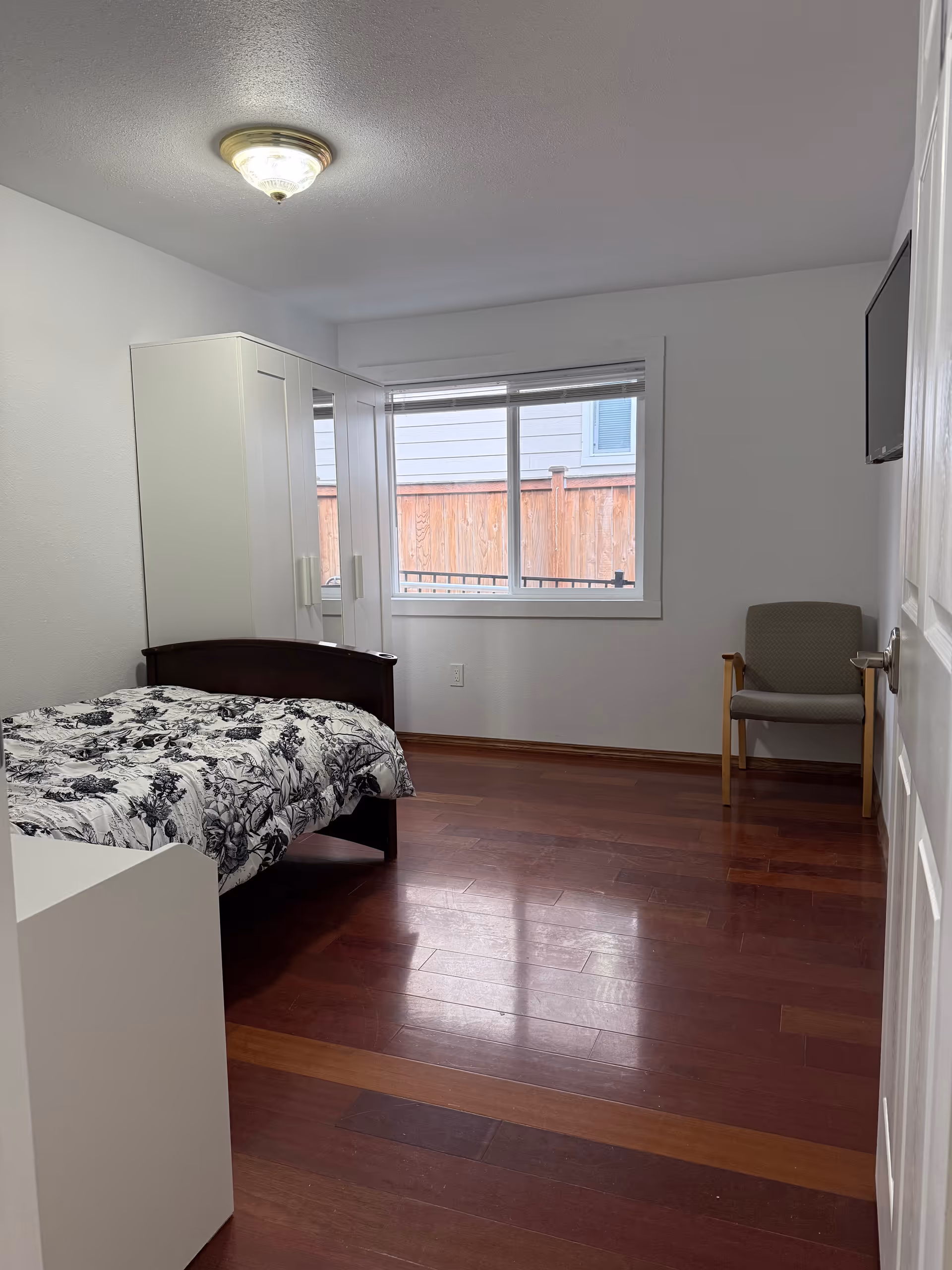 A simple bedroom with a bed, white wardrobe, chair, and window.