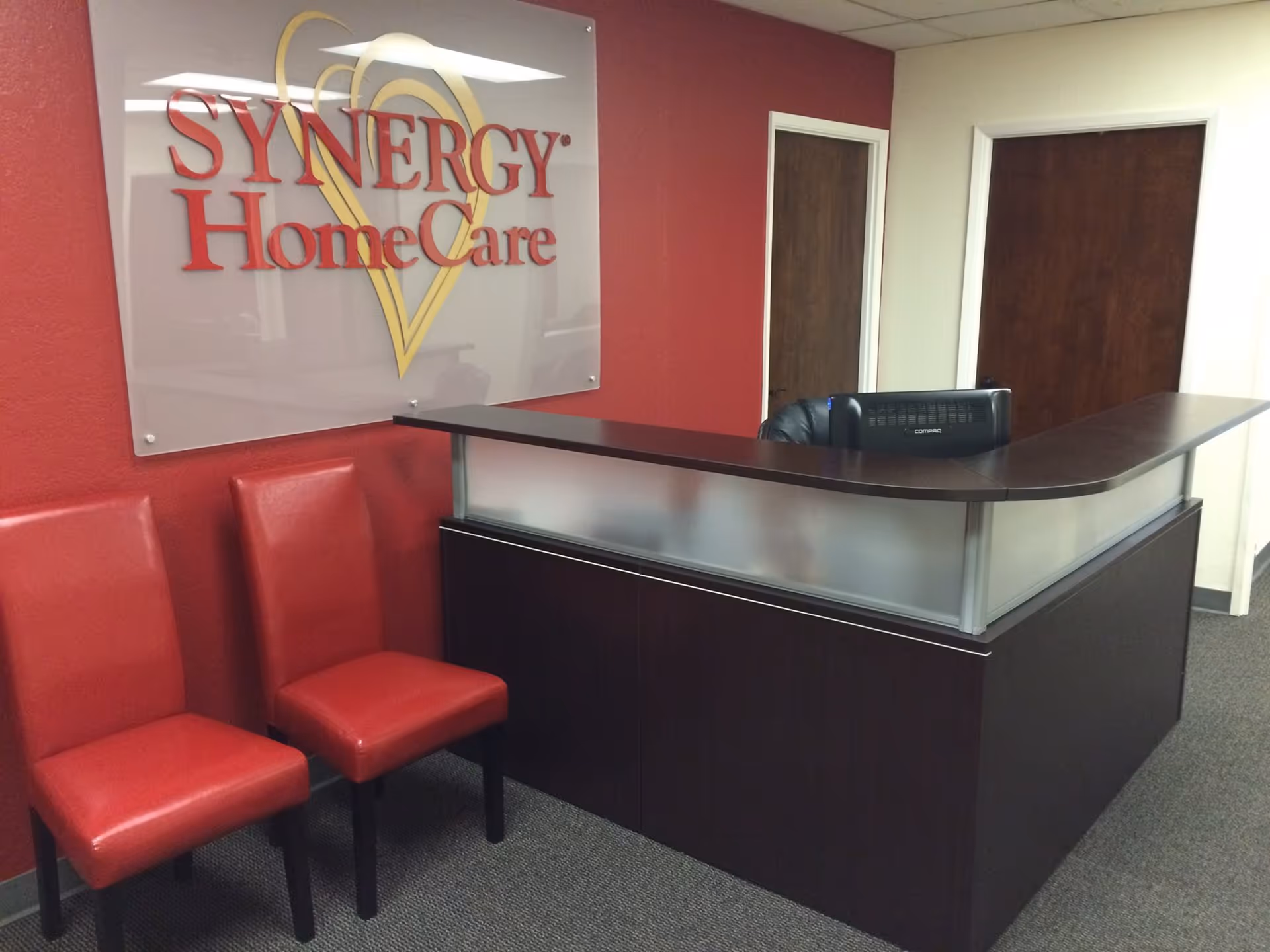 Reception desk with frosted glass panels, red chairs, and a Synergy HomeCare sign.