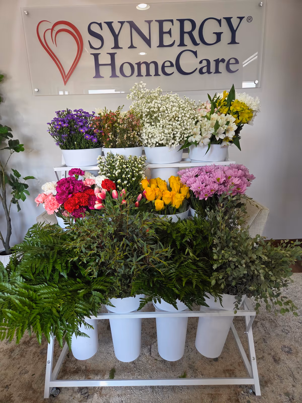 Indoors, a SYNERGY HomeCare sign sits above a colorful flower display in white pots.