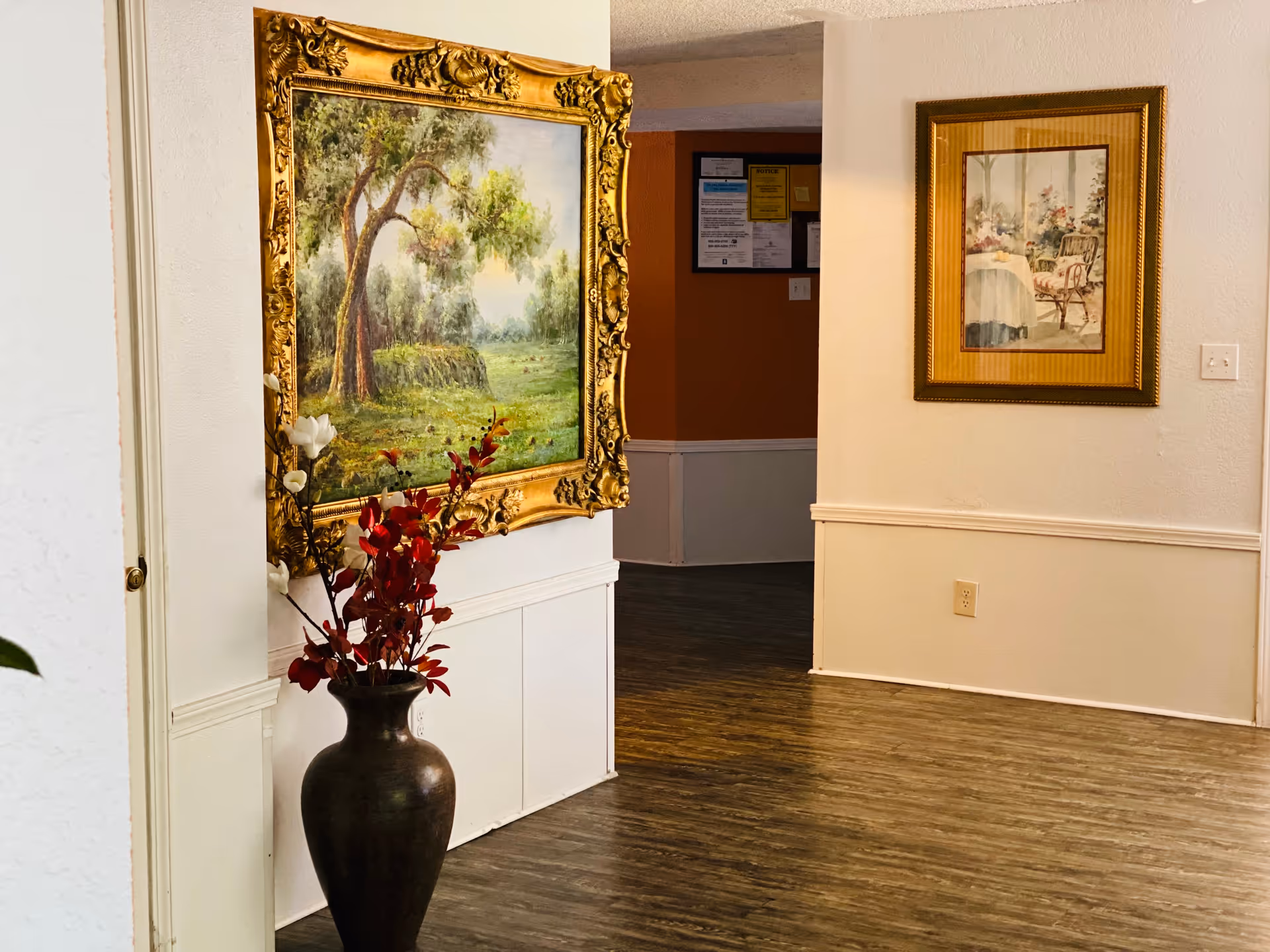 Interior hallway of a senior living facility with framed paintings and a large vase.