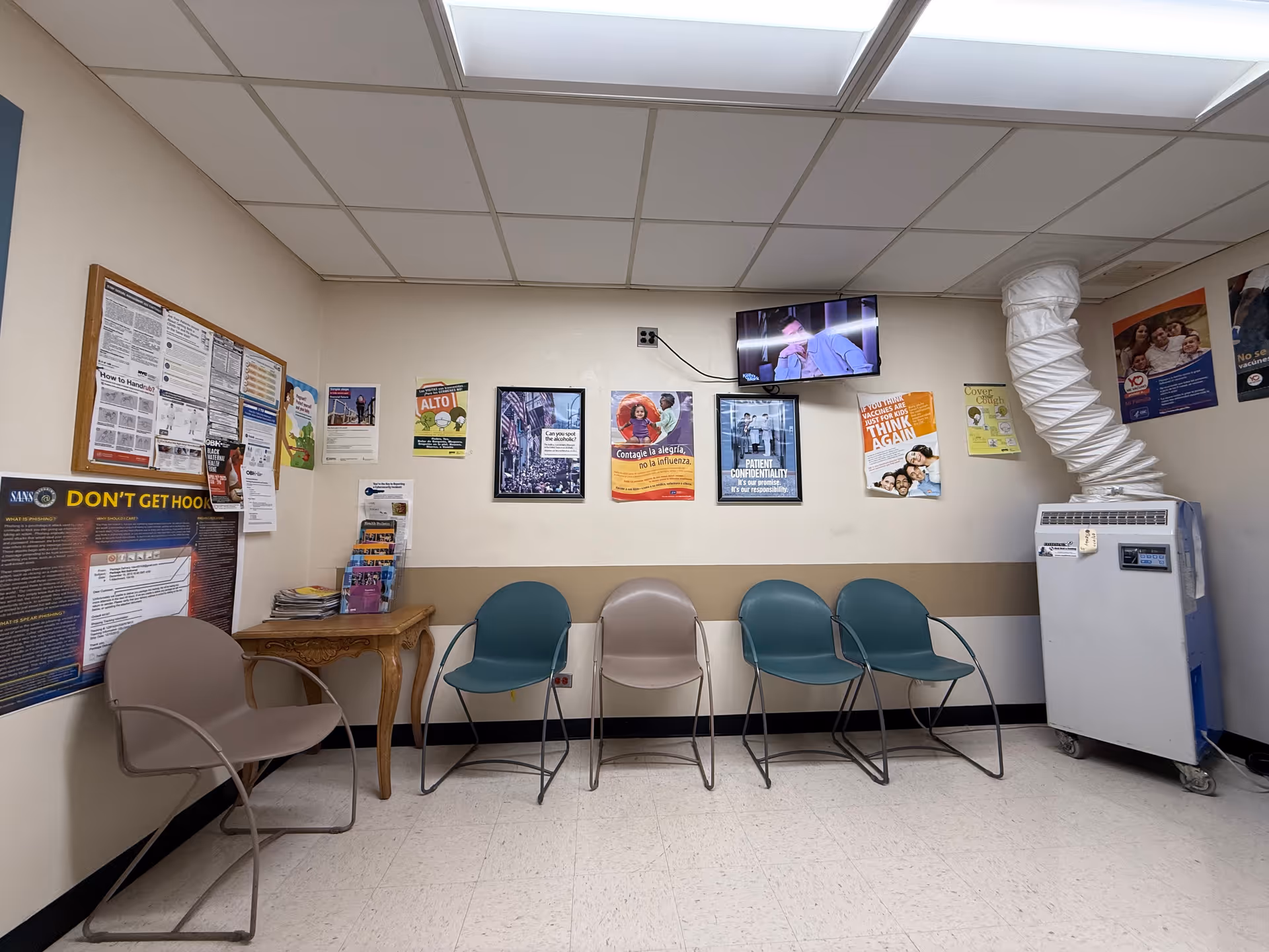 Waiting area with blue and beige chairs, posters, and a vending machine in a hospital hallway.