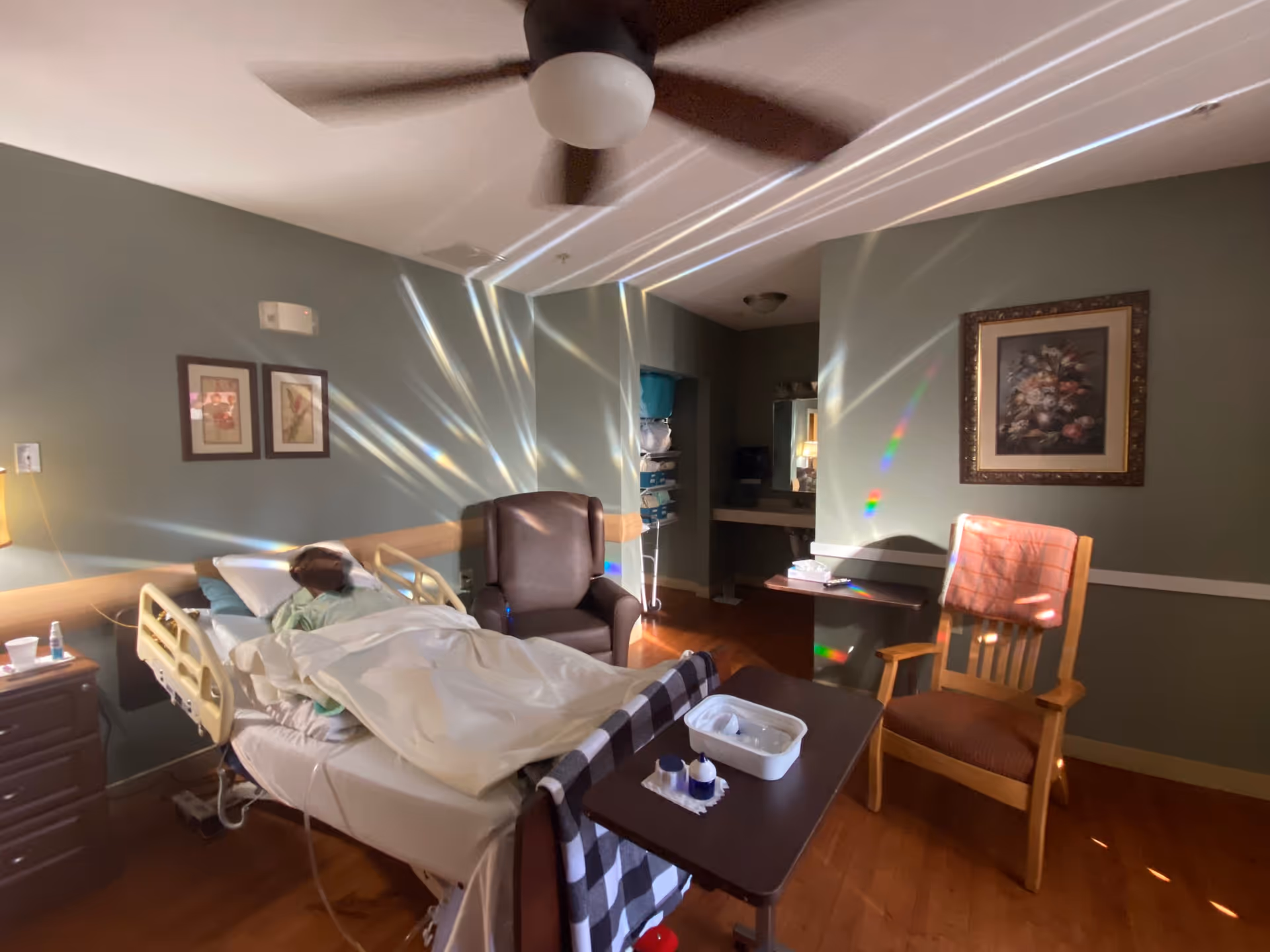 Patient lies in a hospital bed in a dim hospice room with chairs and desk.