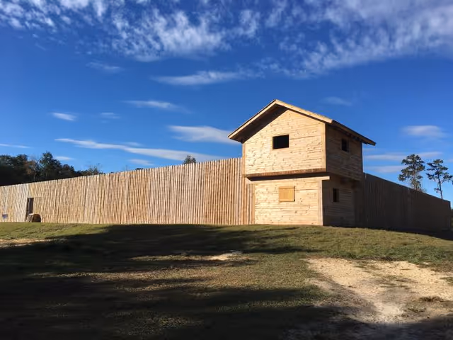 Tall wooden fence with a small two-story stone-like structure under a clear blue sky.