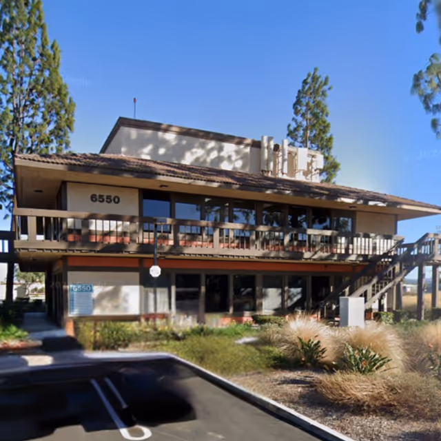 Two-story building with exterior balconies, stairs, landscaping, and a parking lot under a blue sky.