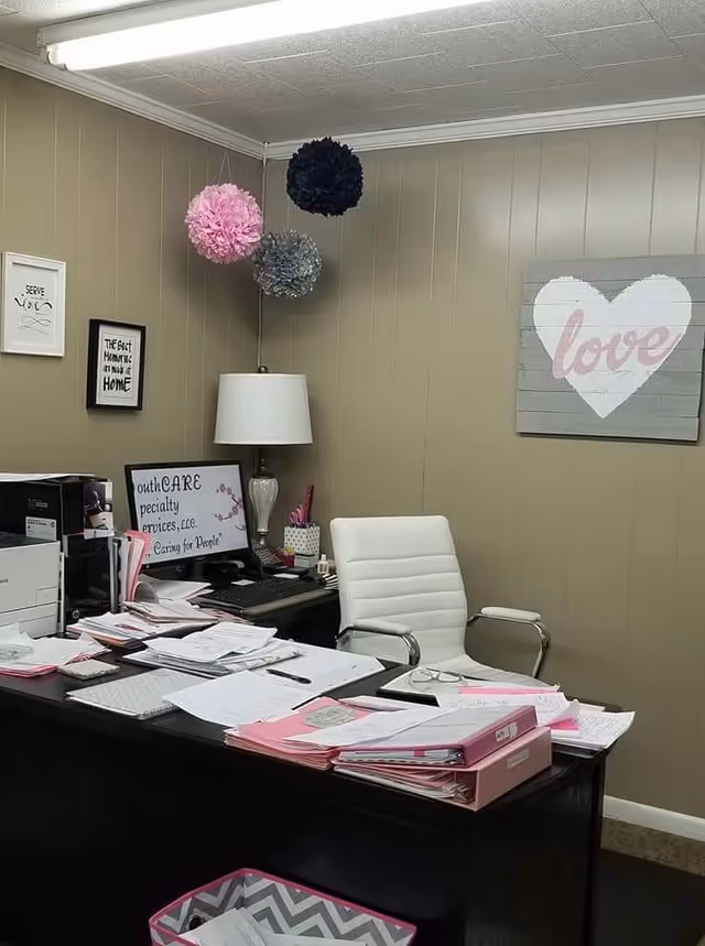 Office desk in a beige-walled facility with papers, pink folders, and a white desk chair.