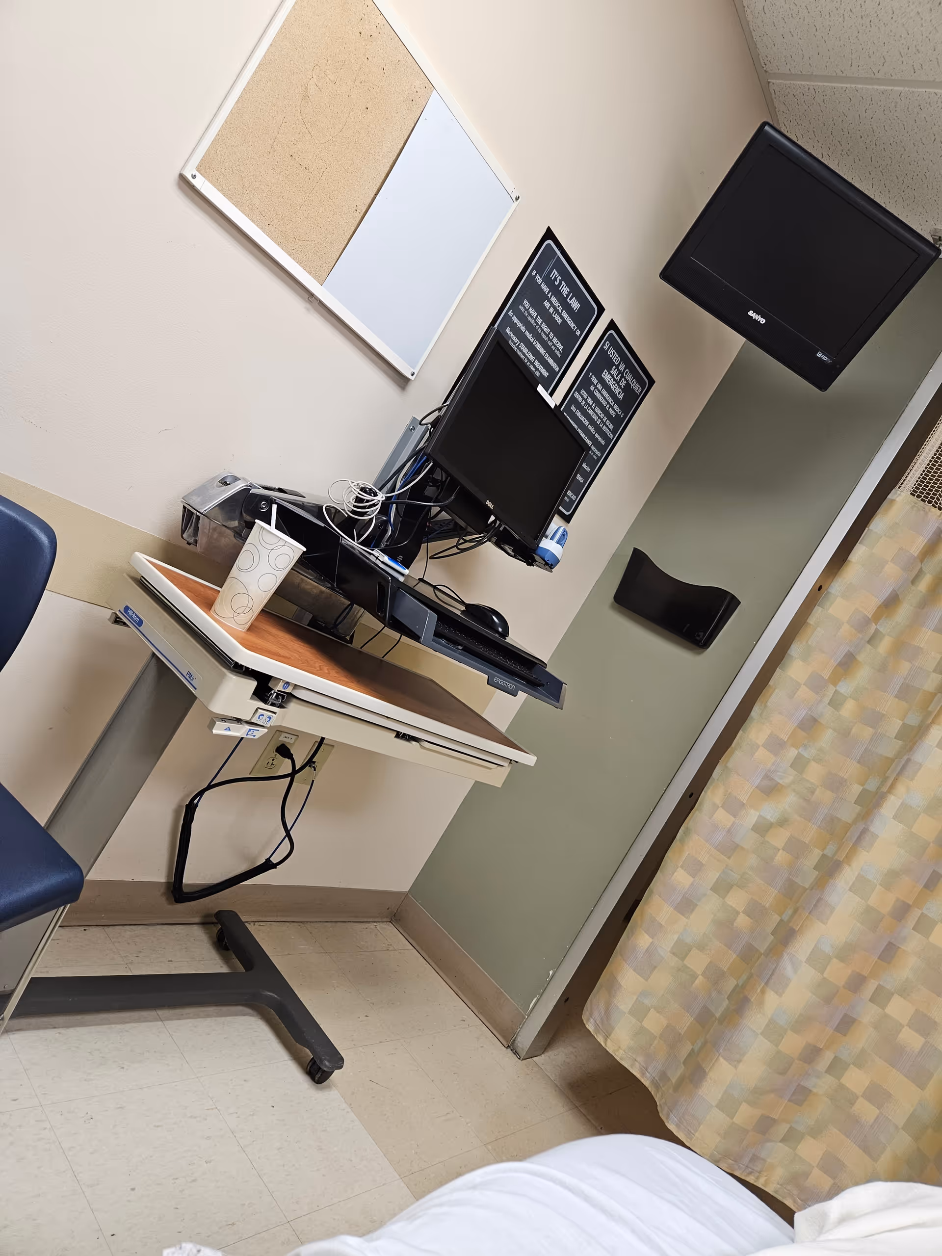 Hospital room workstation with computer monitors, cables, a paper cup, and a bed.