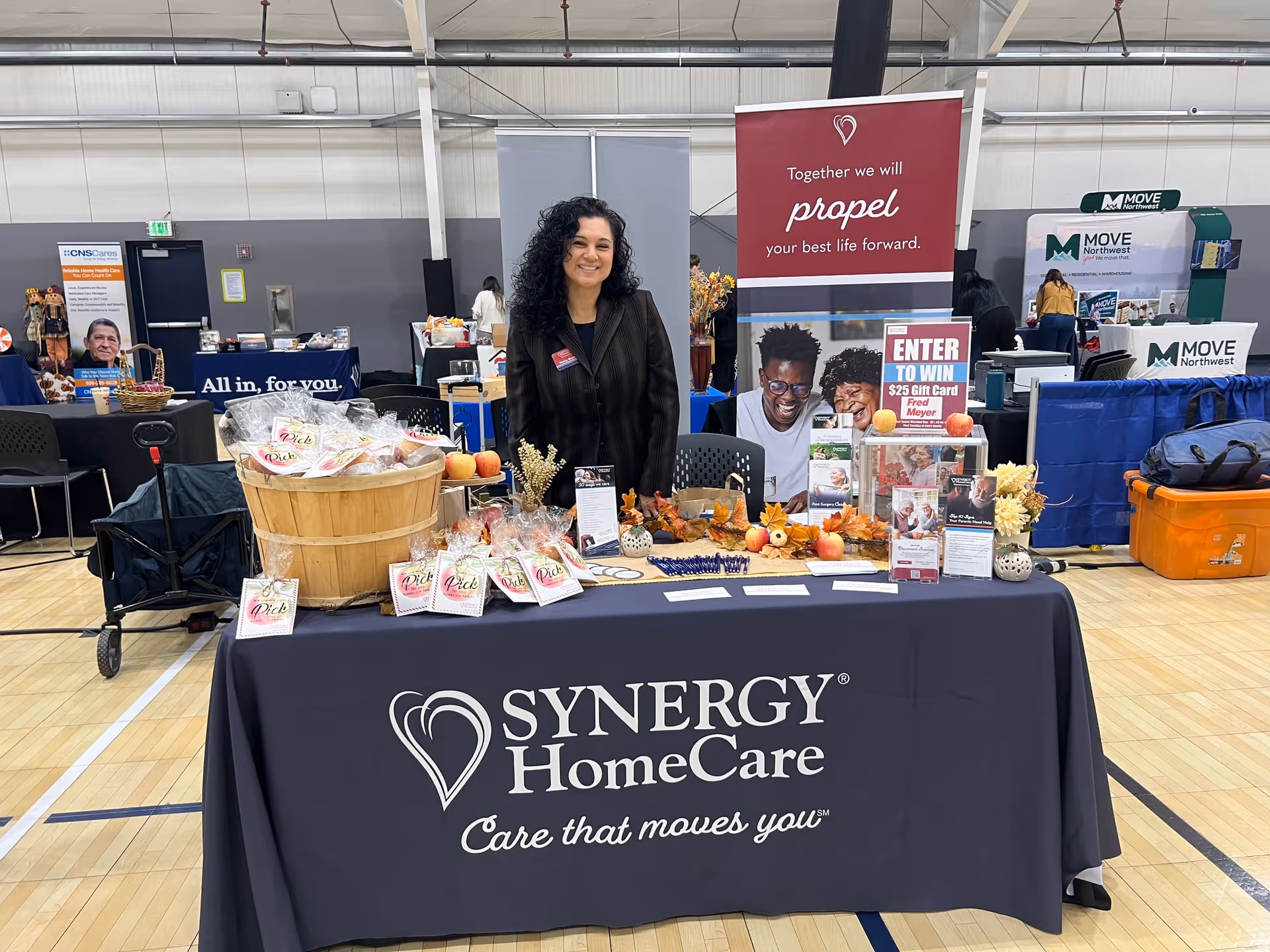 A SYNERGY HomeCare booth with a smiling woman at an indoor expo table.