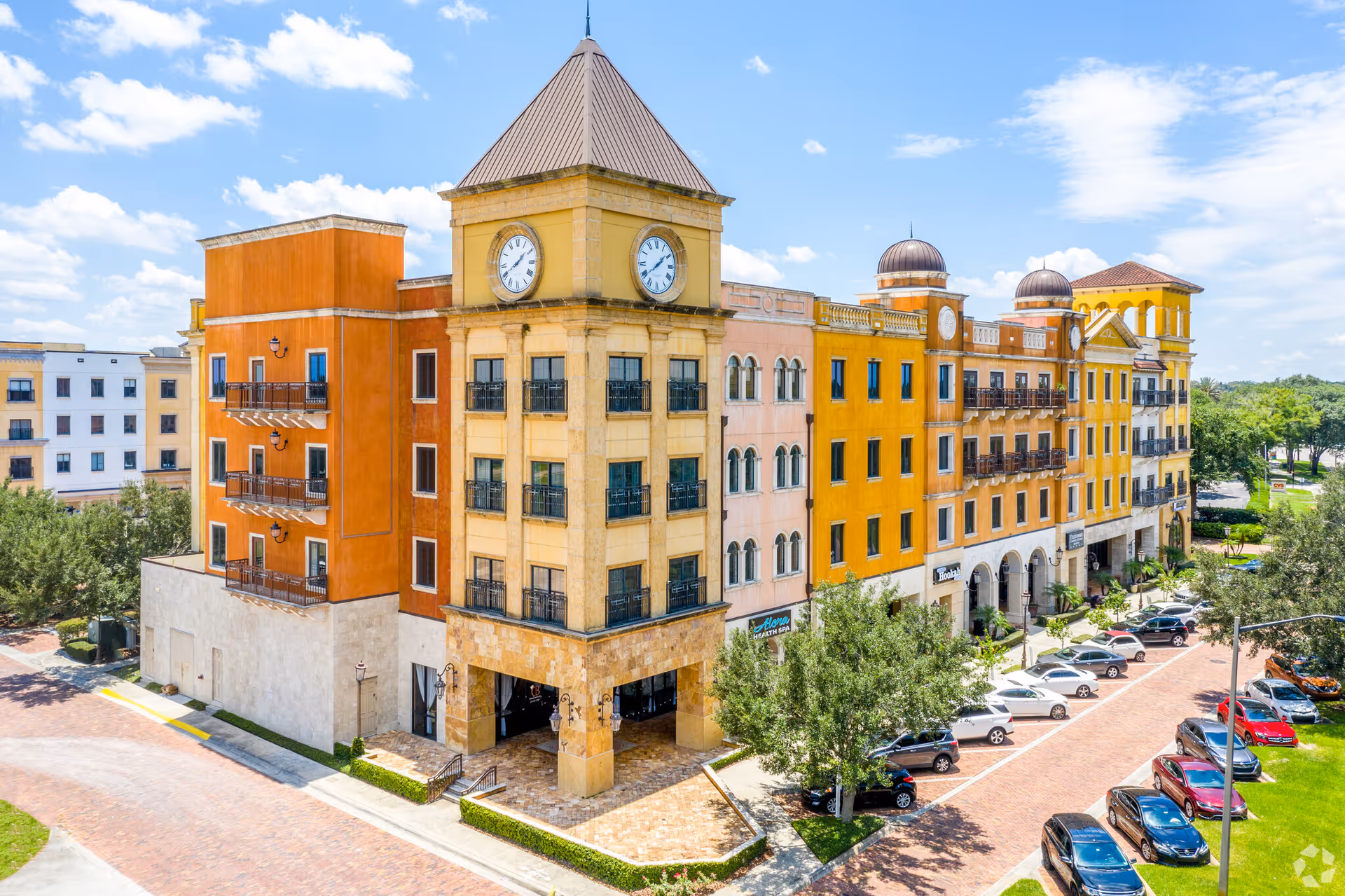 Colorful multi-story senior living building with clock towers, parked cars, and a bright blue sky.
