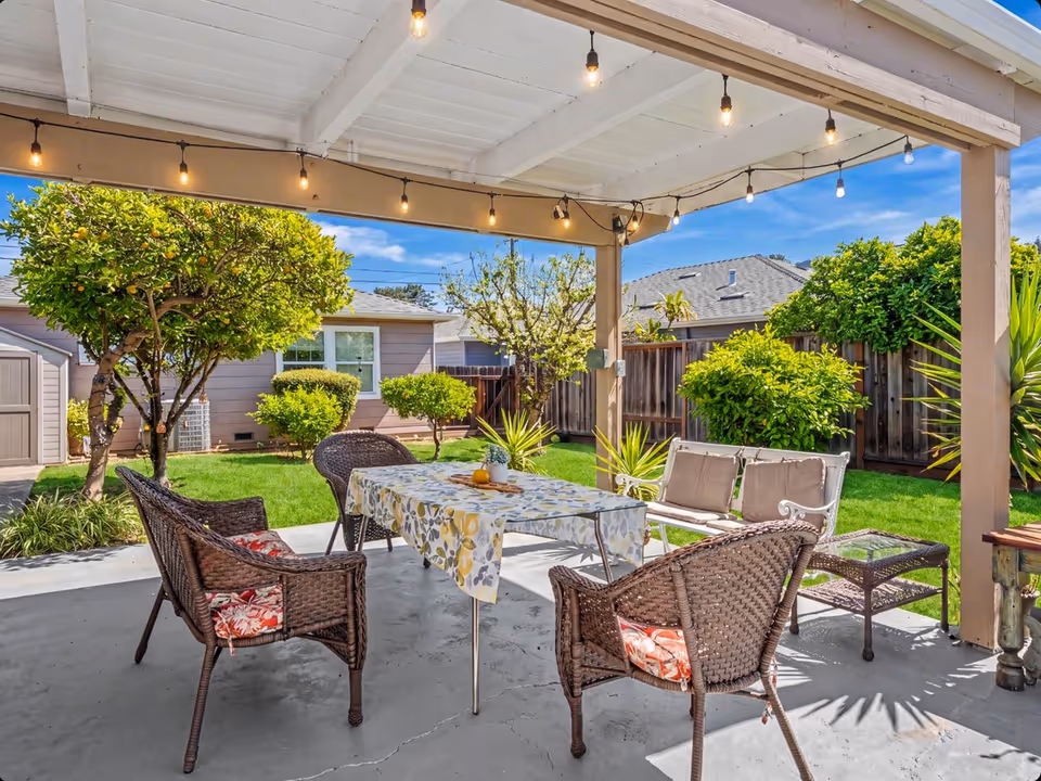 Covered outdoor patio with string lights, a table, and wicker chairs in a sunny backyard.