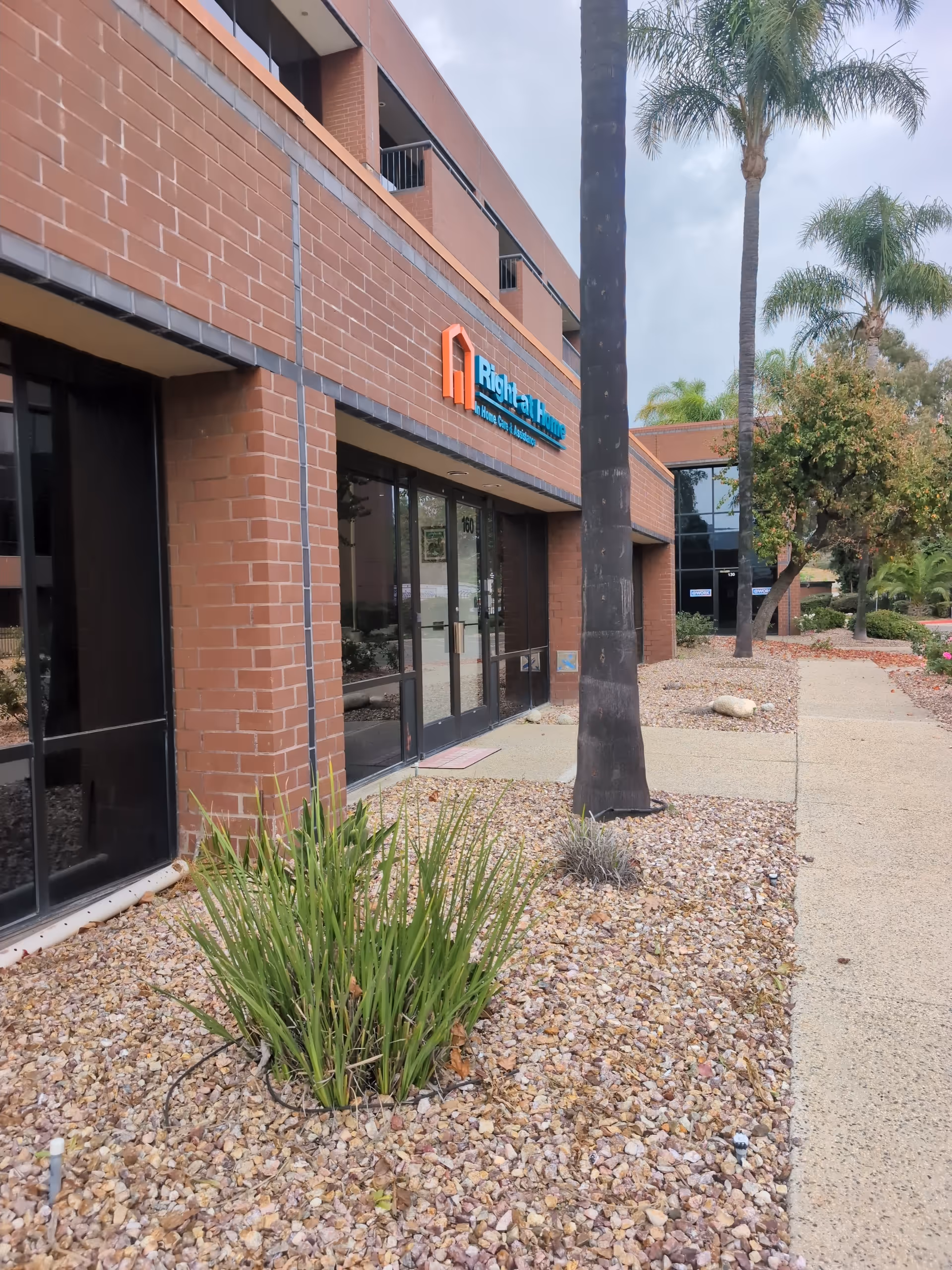 Exterior view of Right at Home facility with brick walls, glass entrance, and palm trees.