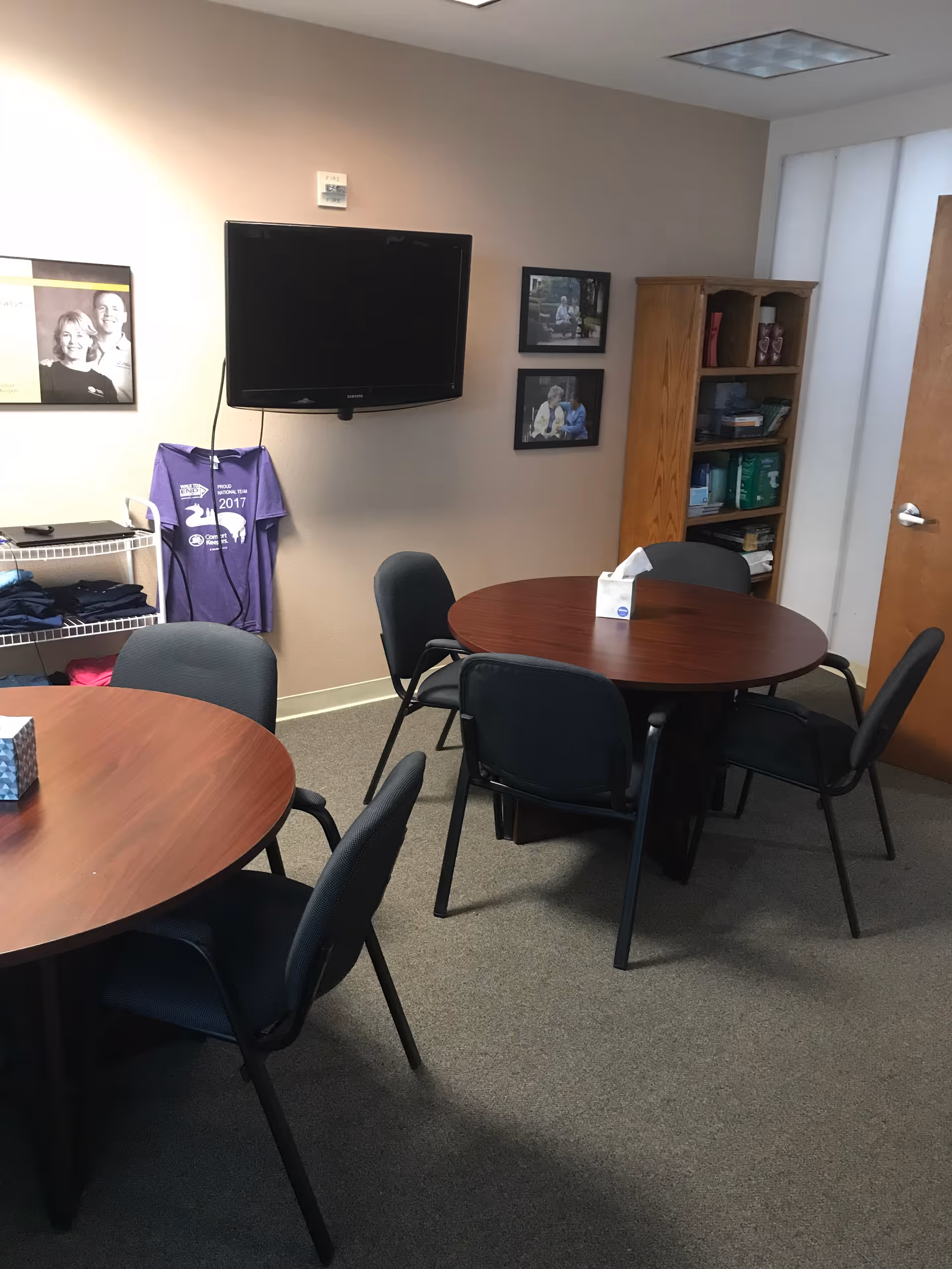 Common room with round tables, chairs, a wall-mounted TV, and a wooden bookshelf.