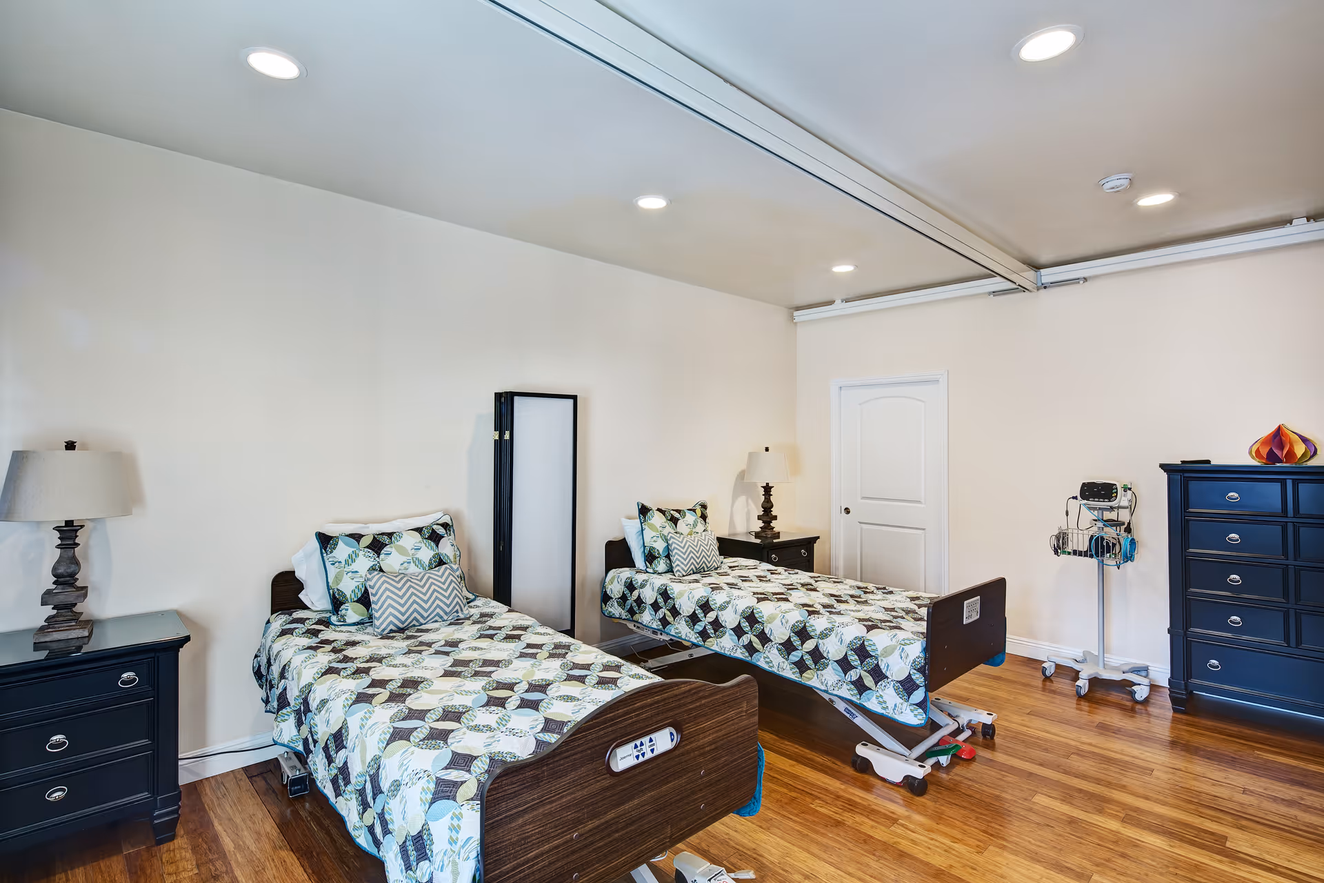 Two hospital-style beds with patterned quilts and lamps in a bright senior living bedroom.