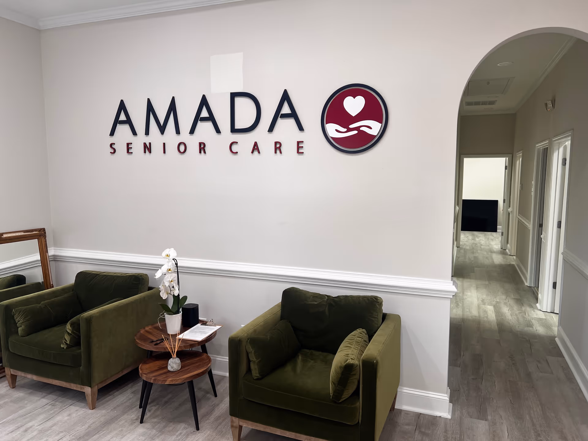 Lobby area with olive-green chairs, small tables, orchid, and Amada Senior Care sign on wall.
