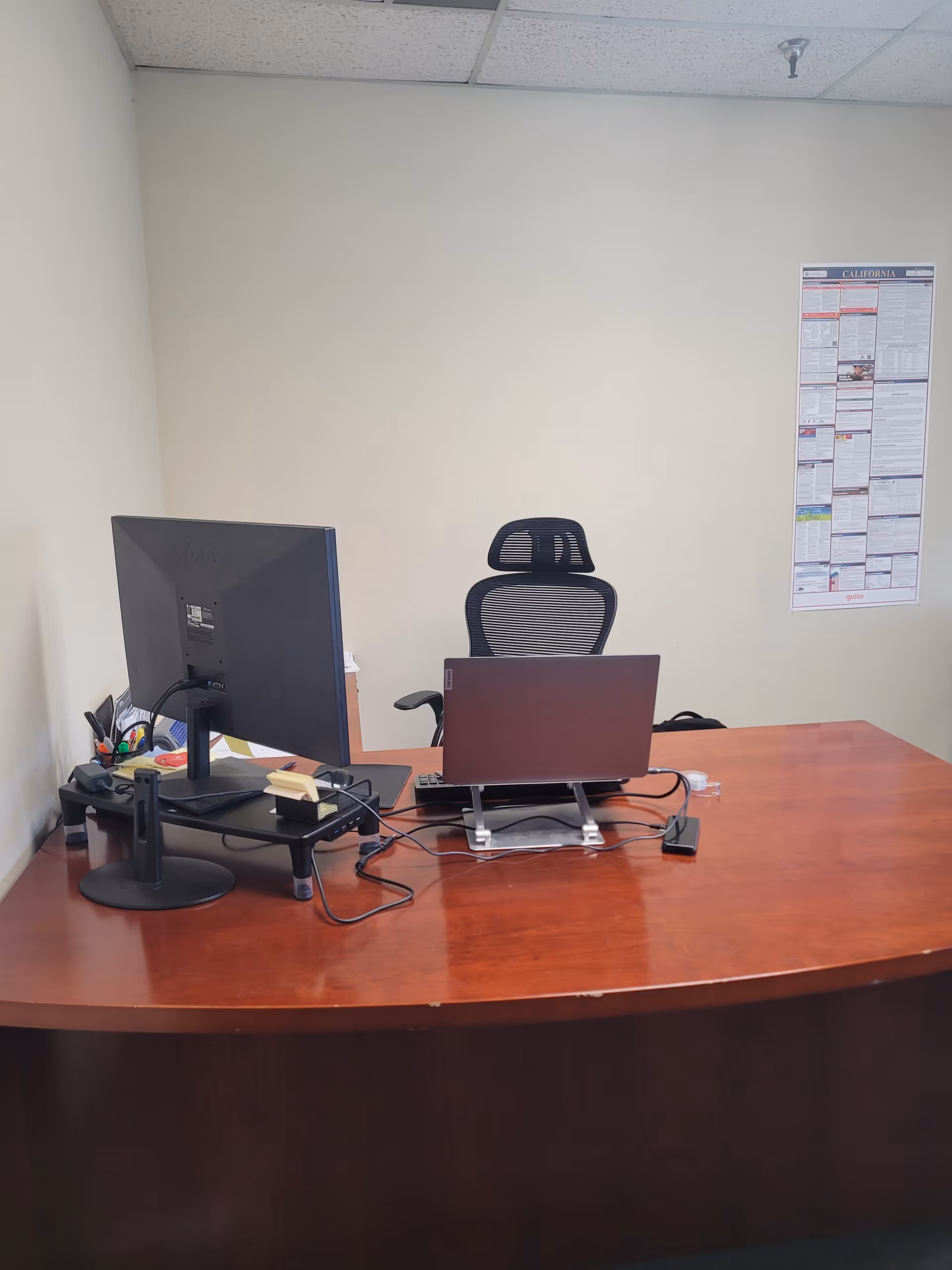Office desk setup with monitor, laptop, cables, and an ergonomic chair against a plain wall.