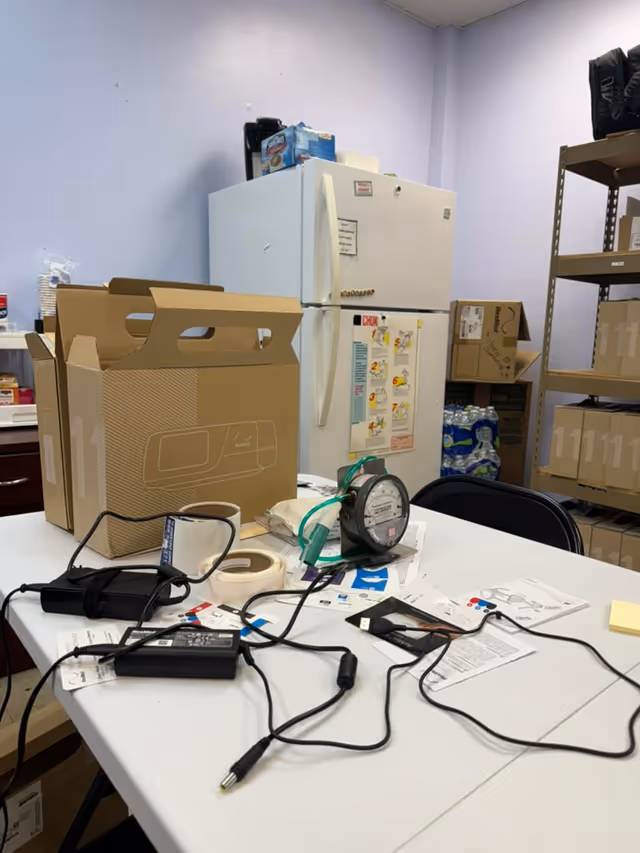 LINCARE supply room clutter with a refrigerator, cardboard boxes, and scattered cords on a table.