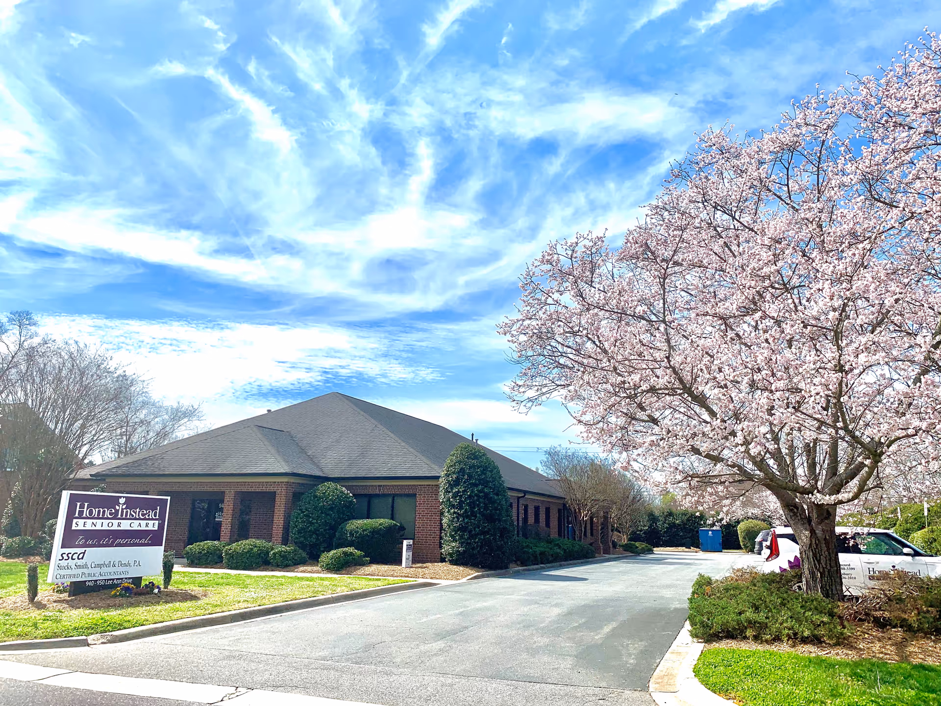 Front exterior of a Home Instead Senior Care building with a blooming tree and blue sky.