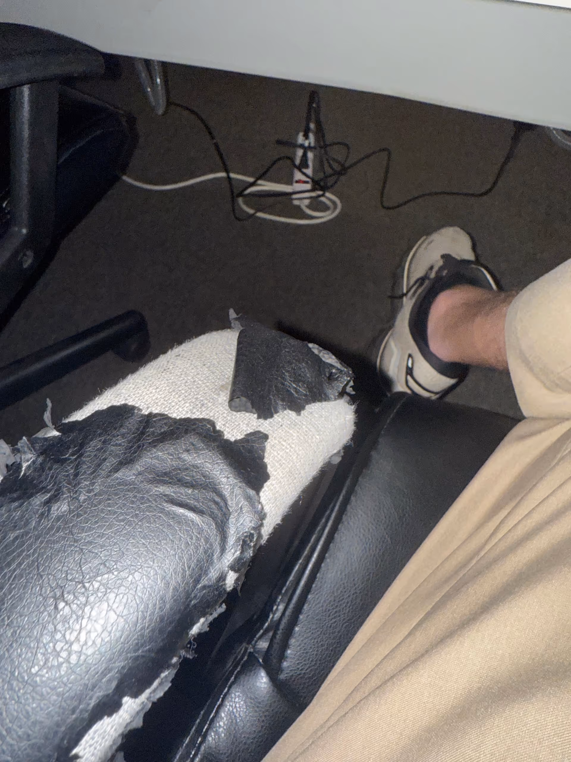 Close-up of torn chair upholstery exposing foam, with a person's leg and sneakers nearby.
