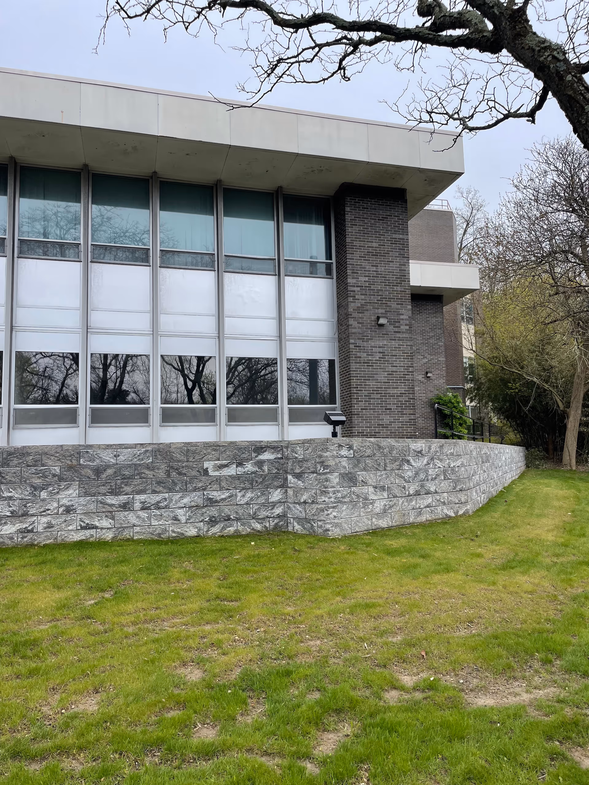Modern brick-and-glass facility facade with large windows, stone retaining wall, and green lawn.