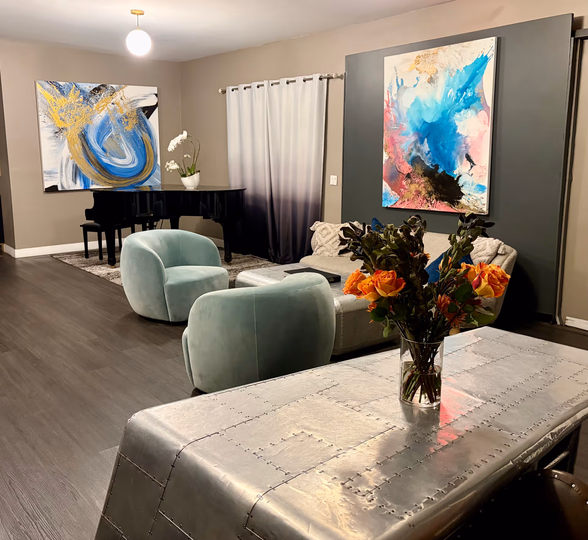 Contemporary lounge with abstract art, a grand piano, blue chairs, and a riveted metal table.