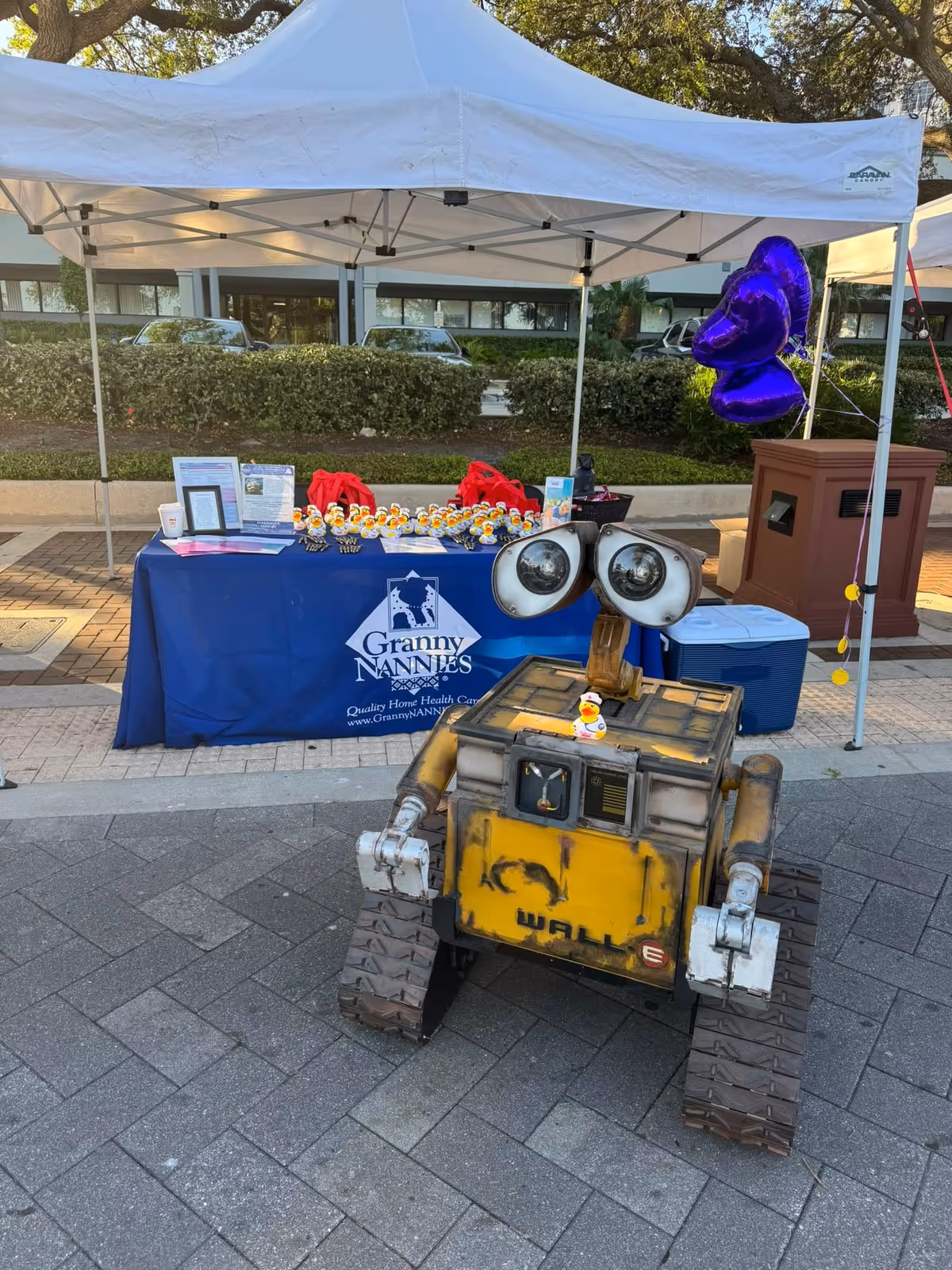Outdoor promotional booth under a white canopy featuring Granny Nannies table and a WALL-E robot.