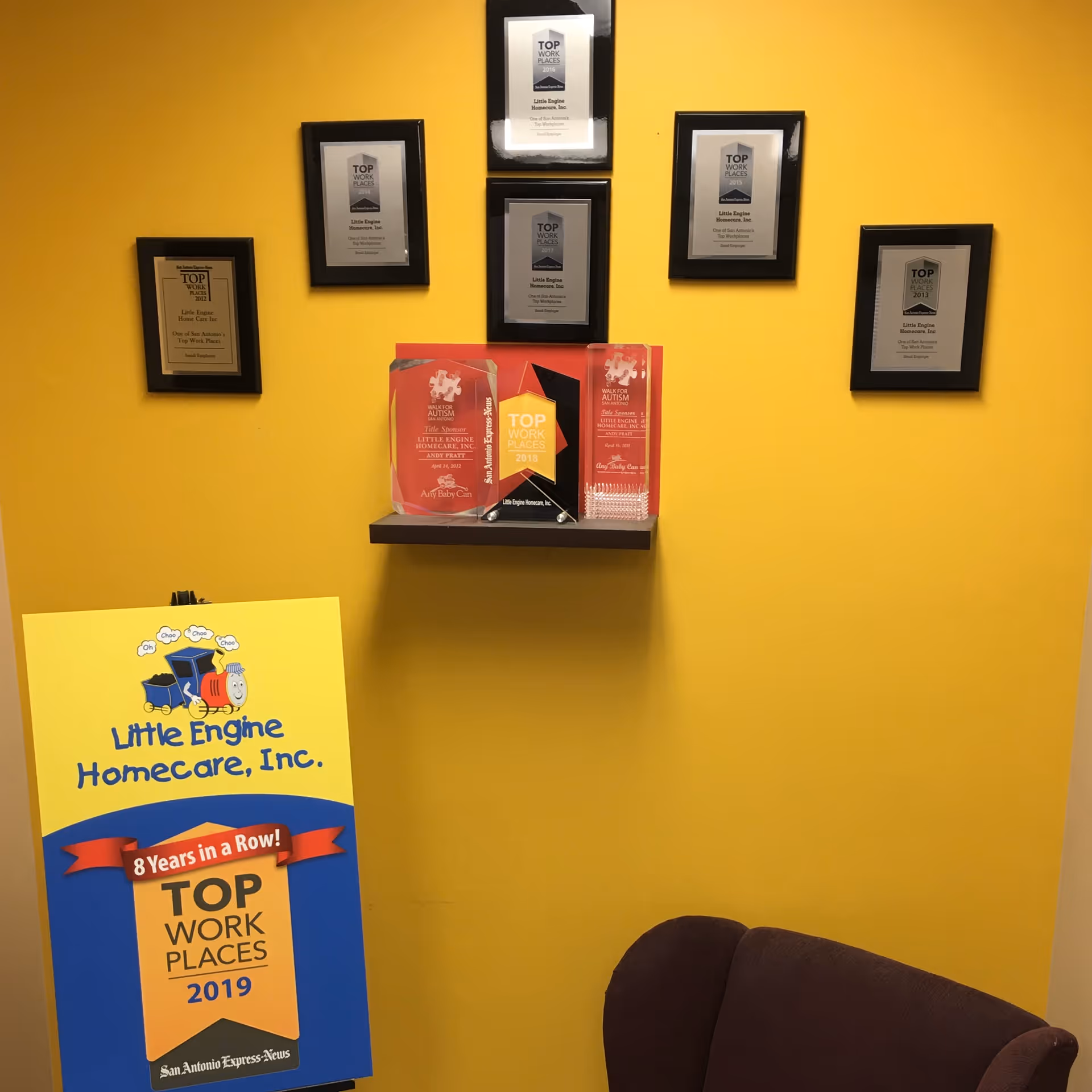 Yellow wall with framed awards, a shelf displaying trophies, and a Little Engine Homecare sign.
