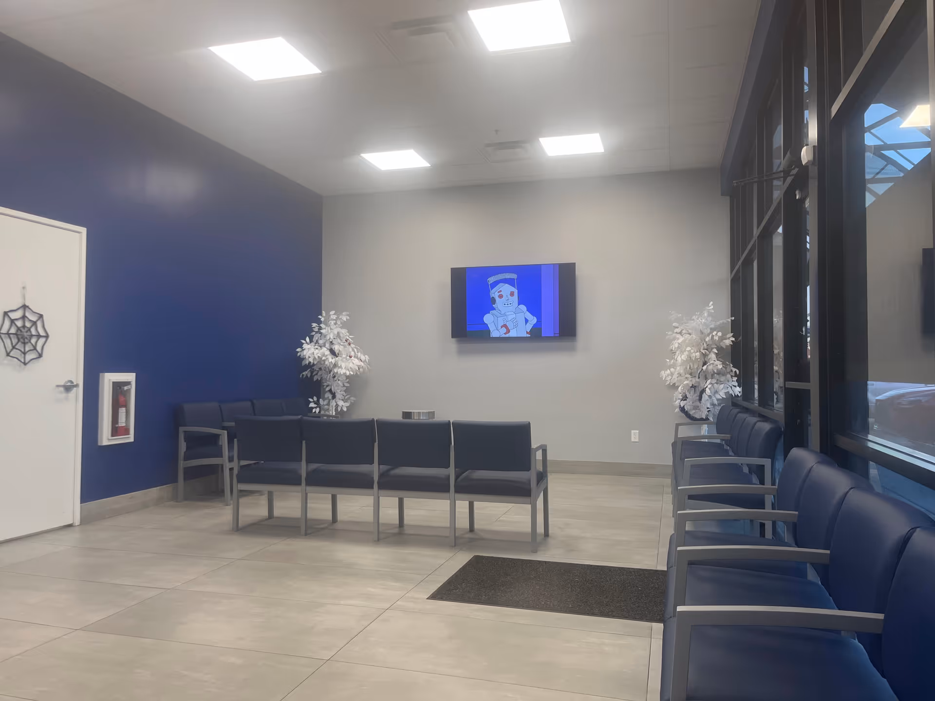 A spacious waiting area with blue accent wall, rows of blue chairs, and a mounted TV.