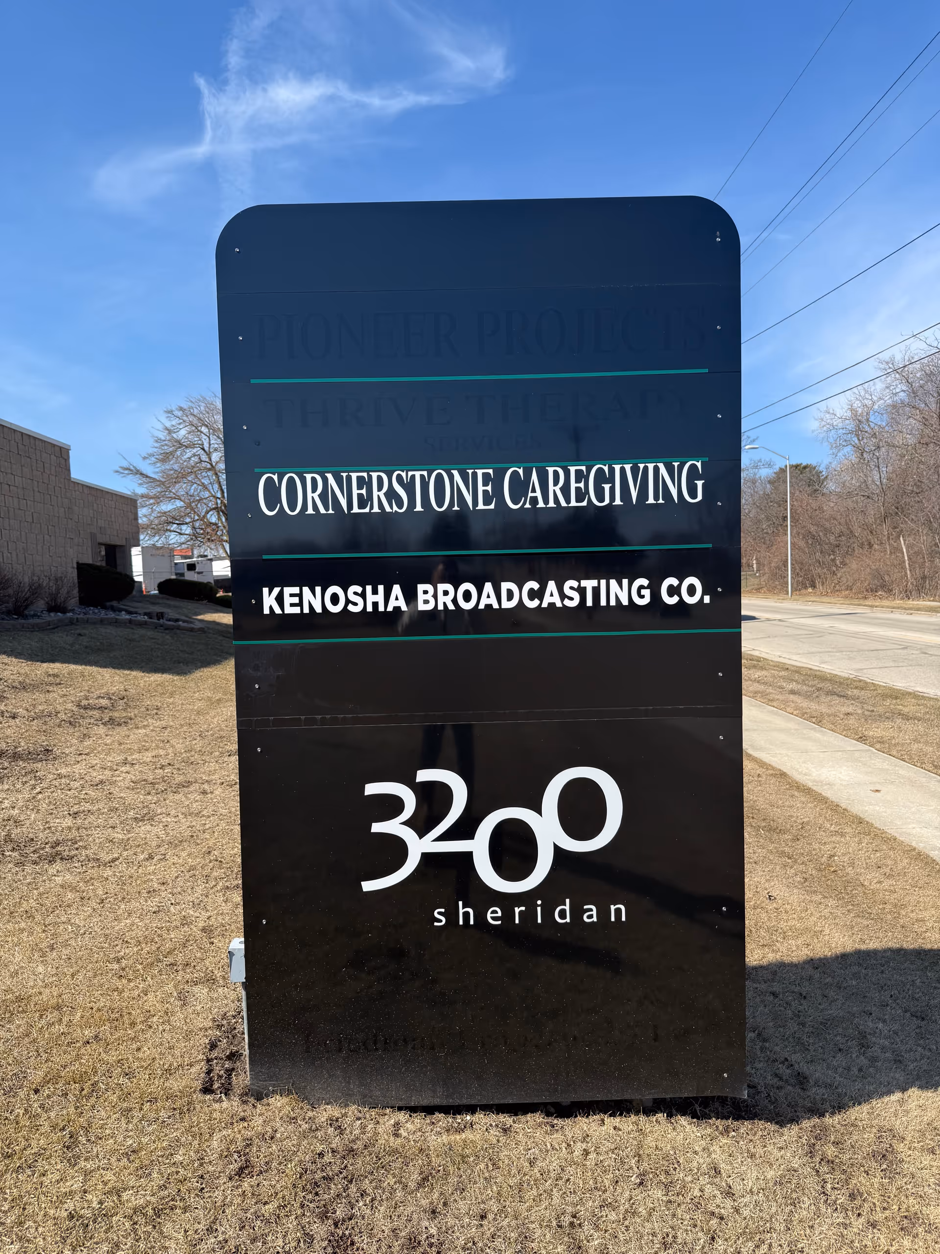 Tall black outdoor sign for Cornerstone Caregiving with address and company logos in a grassy area.