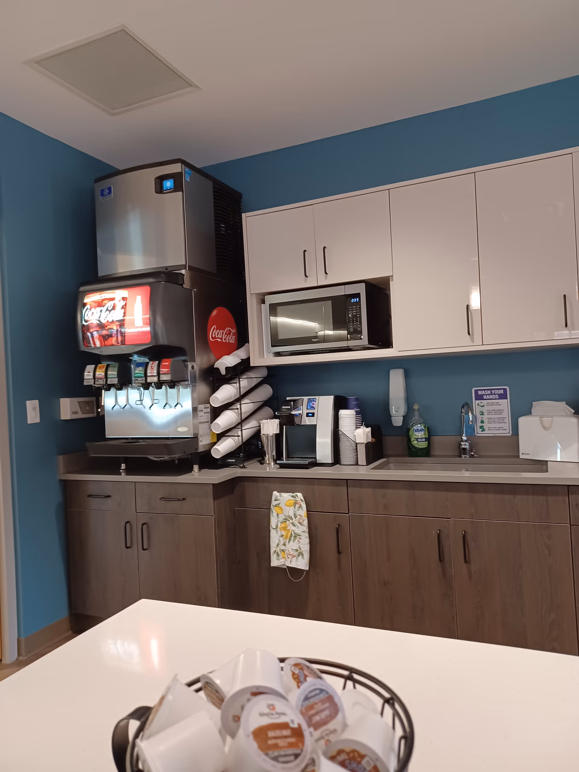 Staff kitchenette with soda dispenser, microwave, cups, and cabinets against teal walls.
